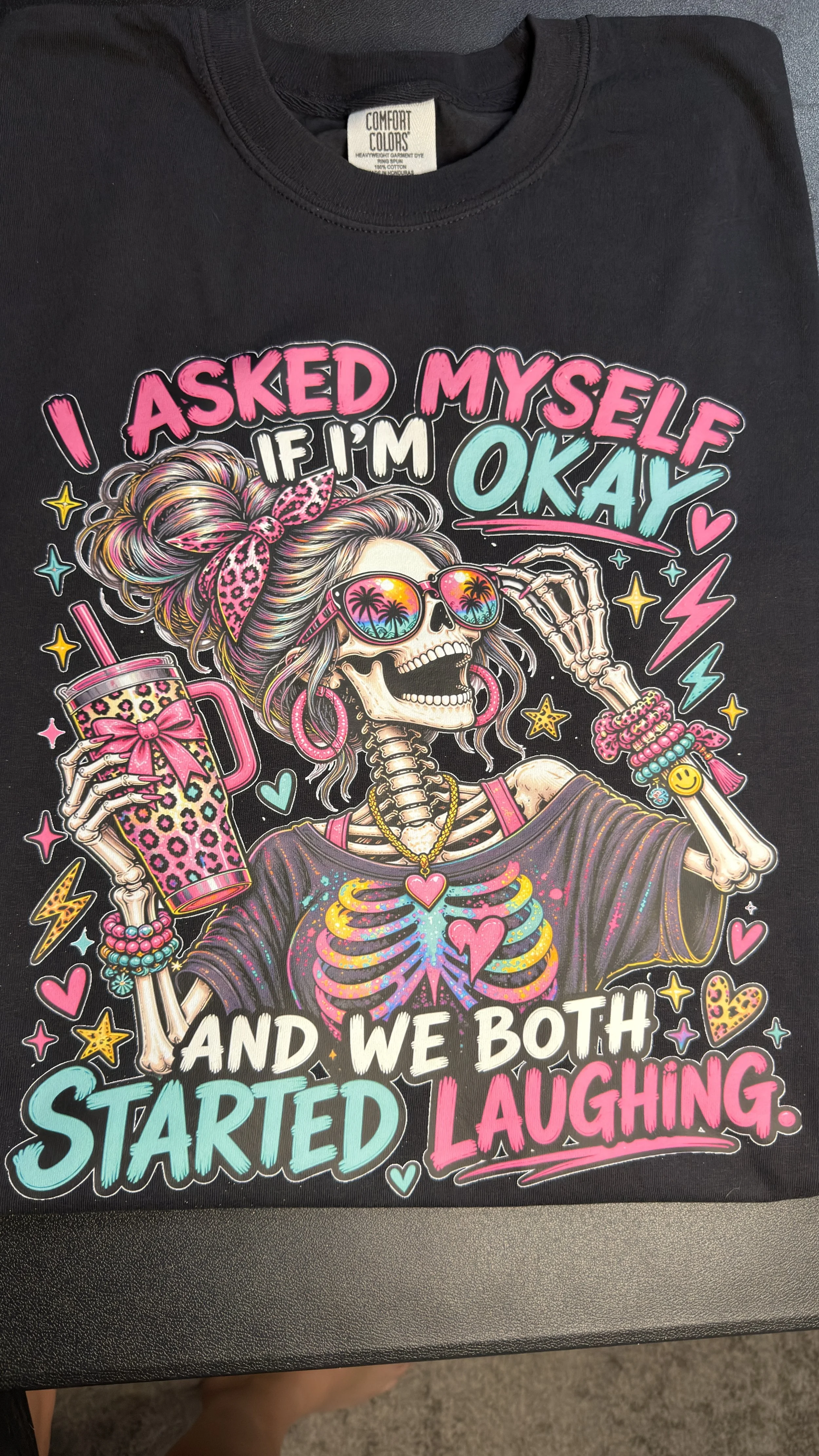 A black T-shirt with colorful graphic design of a skeleton woman wearing sunglasses, a bandana, and jewelry, holding a drink, surrounded by hearts, stars, and lightning bolts. The shirt displays the text 'I asked myself if I'm okay and we both starte