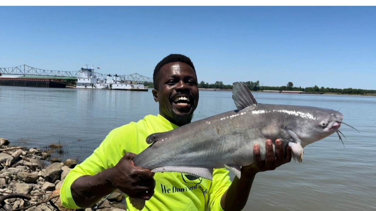 Man in bright yellow shirt holding large catfish by water, smiling, with bridge and ship in background.