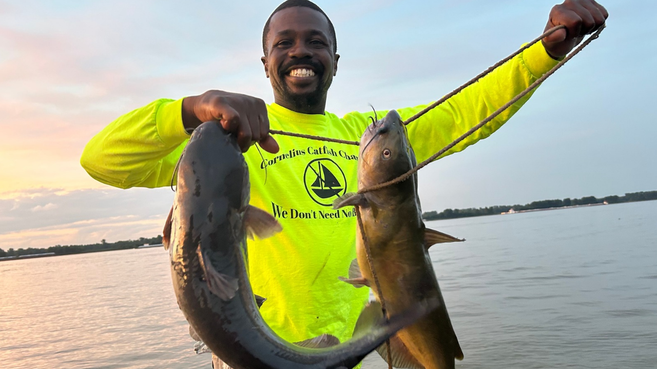 Cornelius Catfish smiling and holding two fish by a stringer while standing near a body of water, with a sunset sky in the background. He is wearing a bright yellow-green long-sleeve shirt with black text and a logo.