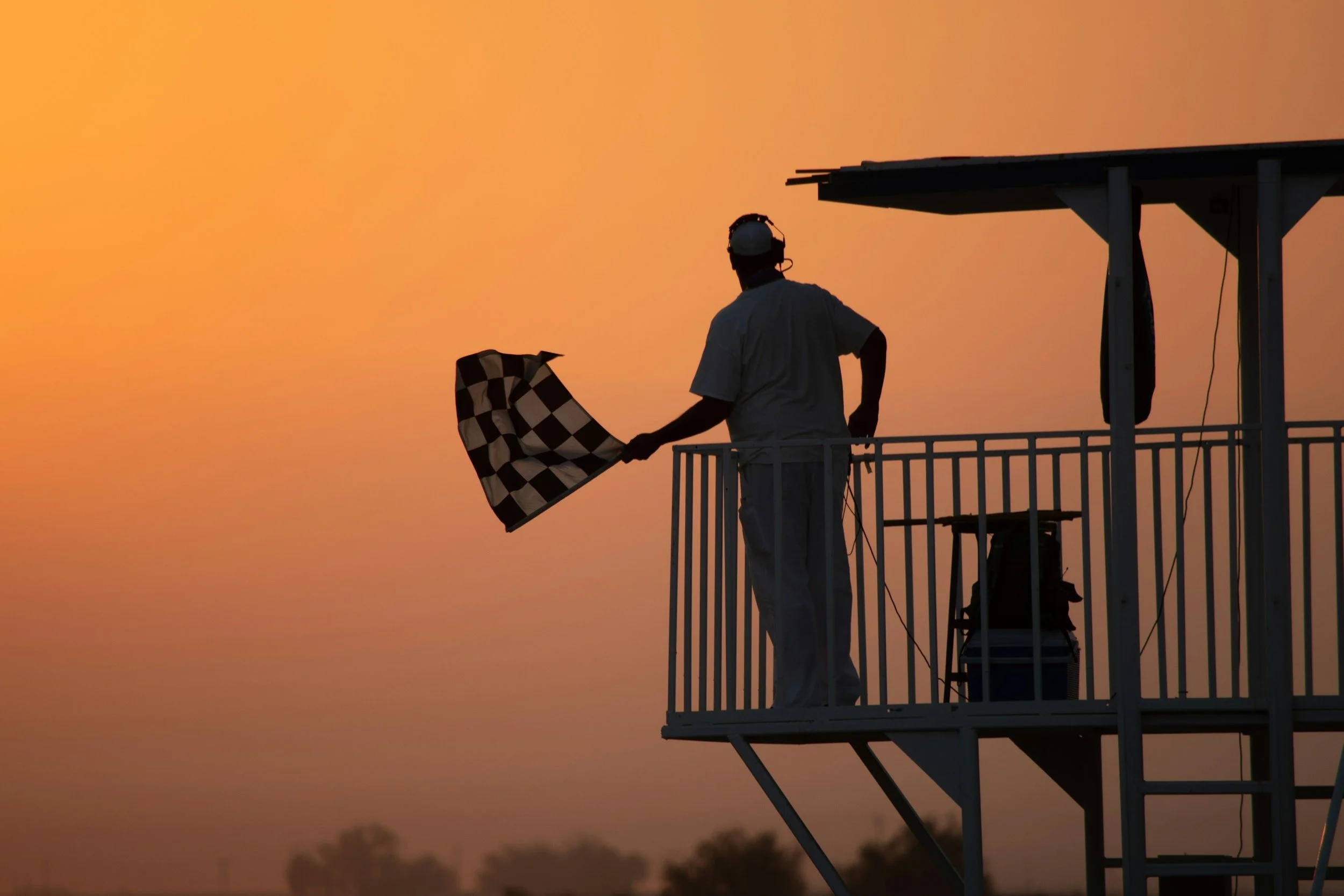 A silhouette of a person standing on an observation deck, holding a checkered flag, with a sunset or sunrise sky in the background.