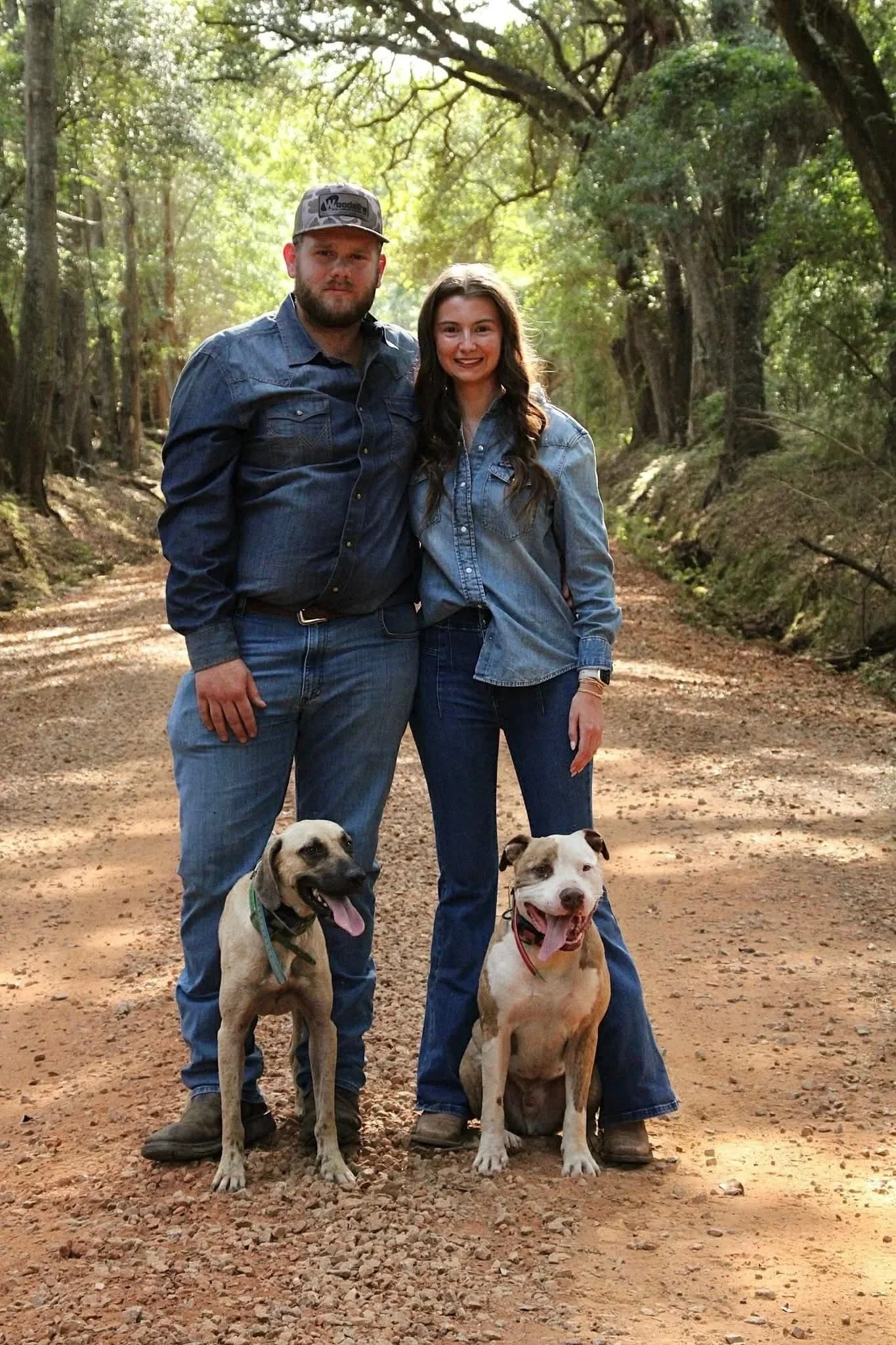 A man and a woman standing side by side on a dirt trail in a forest, with two dogs sitting in front of them. The man is wearing a denim shirt and a cap, and the woman is dressed in a denim jacket and jeans. The dogs are large, one with a tan and white coat, and the other with a gray and white coat, both with their tongues out. Sunlight filters through the trees, creating a peaceful outdoor setting.