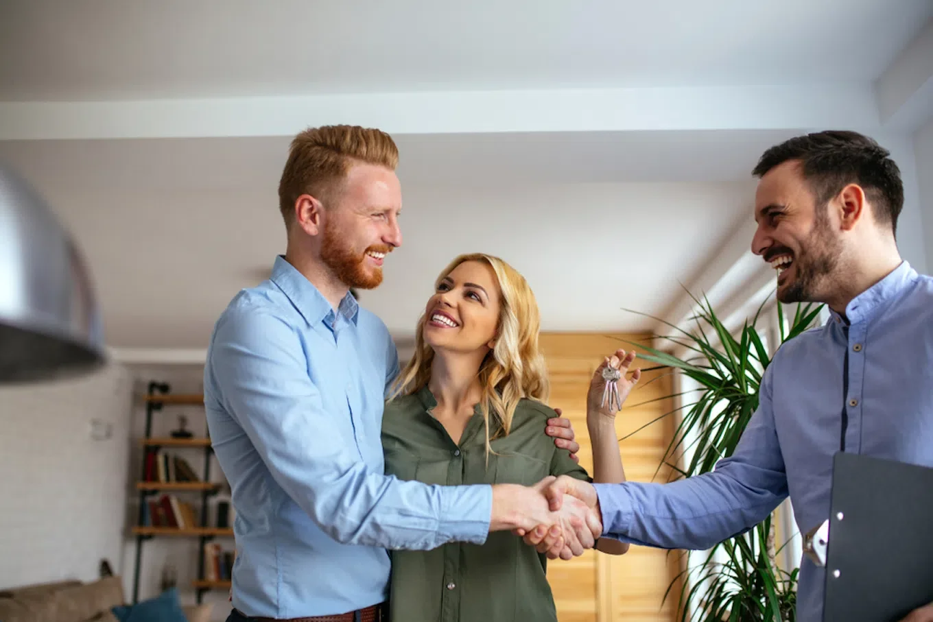 Two men shaking hands in an office, smiling, while a woman stands between them, smiling and holding keys.
