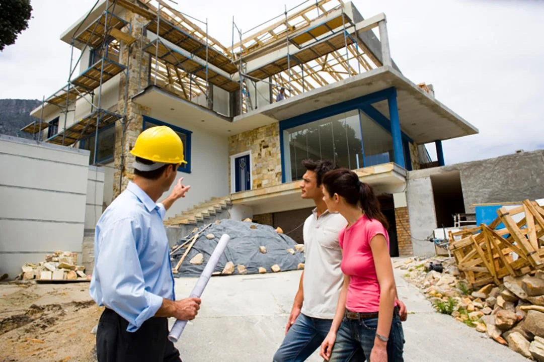 Construction site with a modern house under construction, two men and one woman discussing building plans, one man wearing a yellow safety helmet, scaffolding around the house, construction materials scattered on the ground.