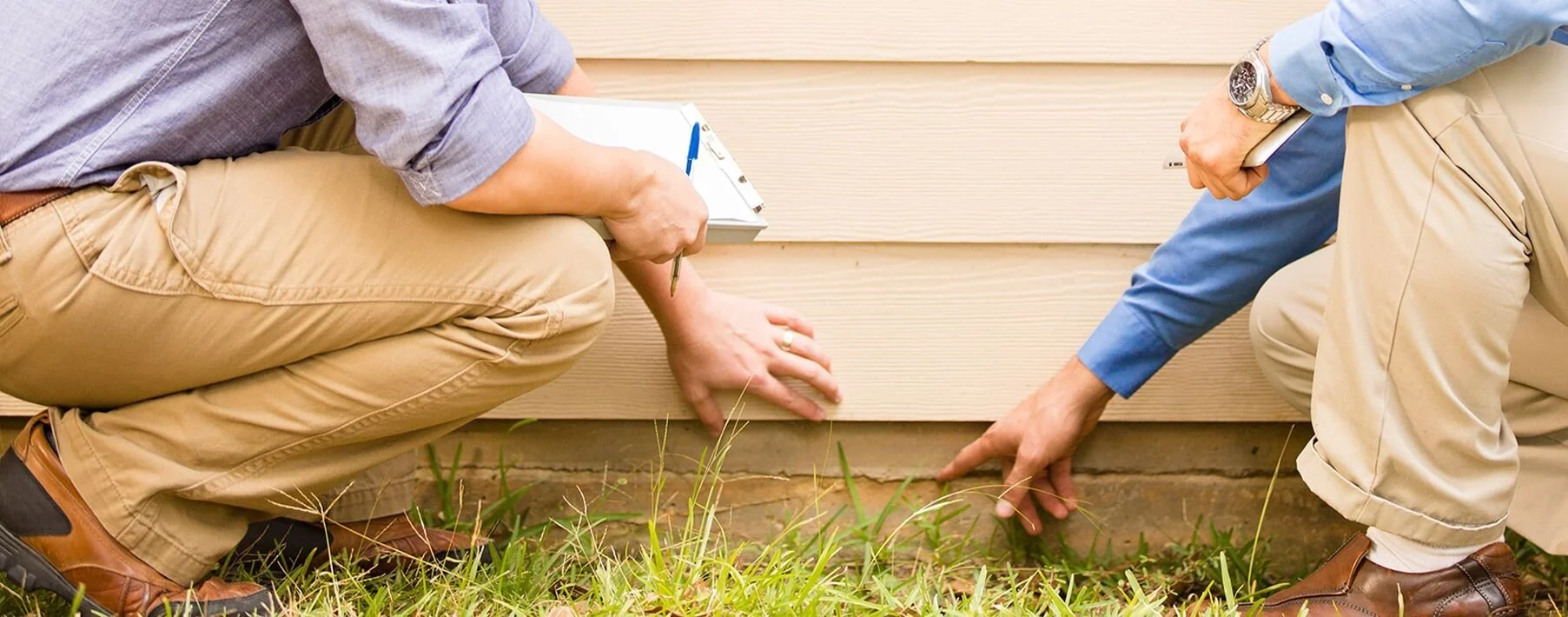 Two men crouch down near the foundation of a house, examining and pointing to the ground and bottom siding with grass nearby.