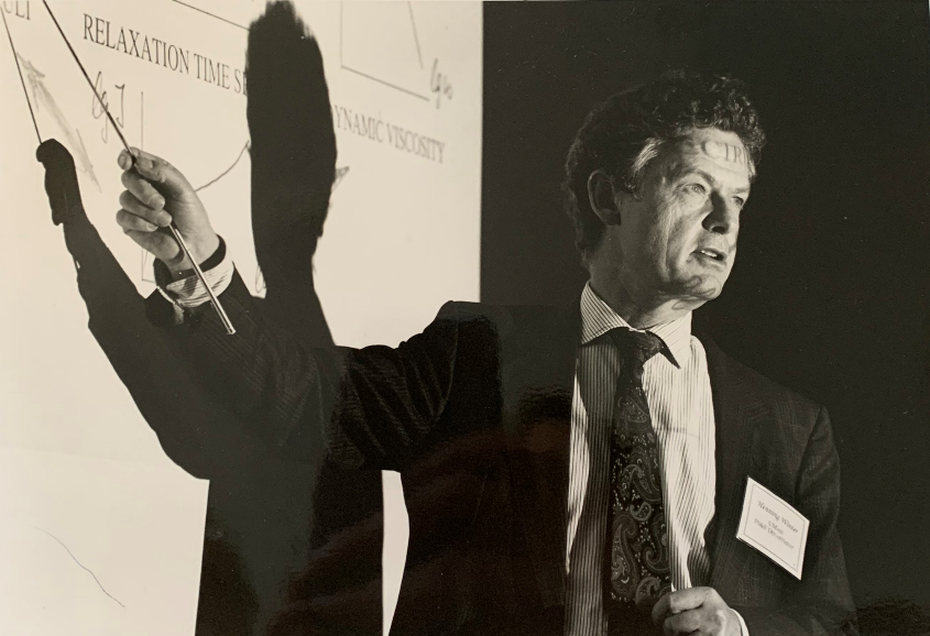 A man in a suit and tie giving a presentation with a projected slide. He is pointing to the slide with a stick, casting a shadow on it, while looking to the side.