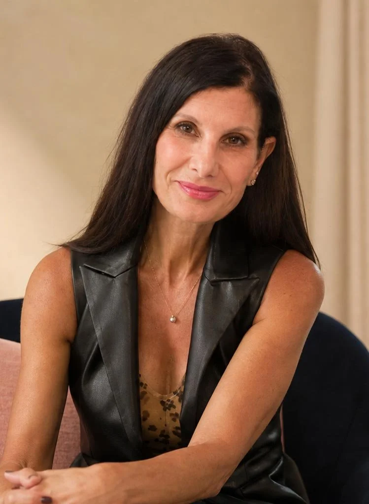 A woman with long dark hair, wearing a sleeveless leather vest and a necklace with a small pendant, sitting on a couch and smiling at the camera.