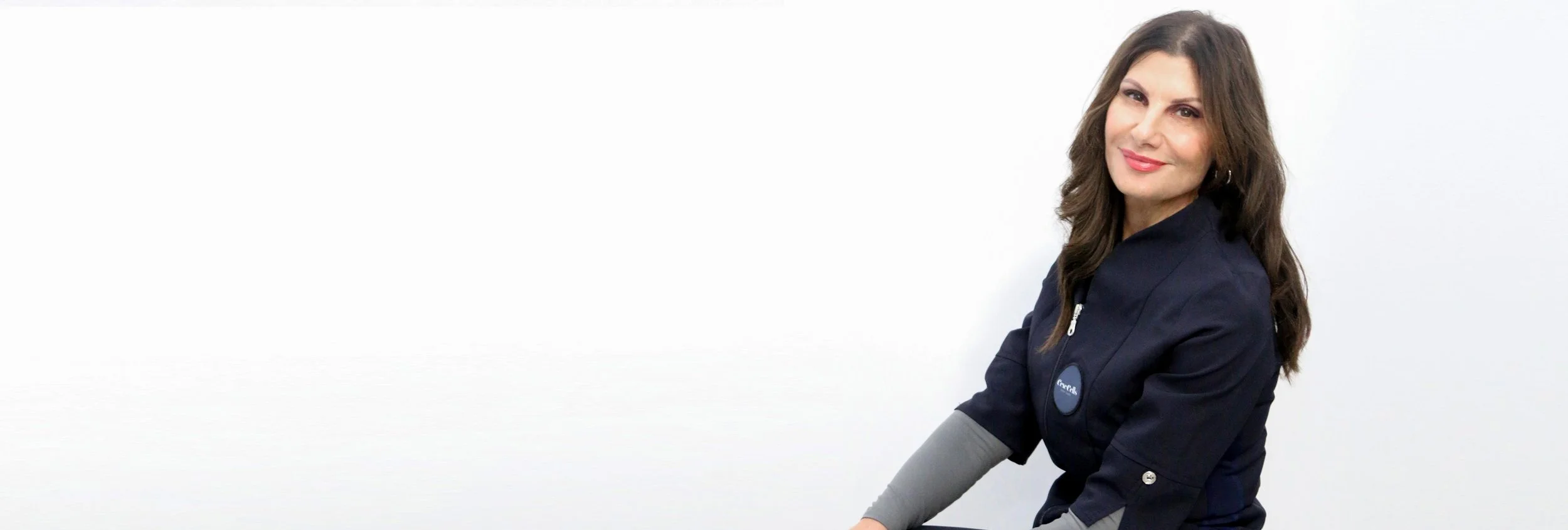Woman with long brown hair wearing a dark blue jacket, sitting and smiling against a plain white background.