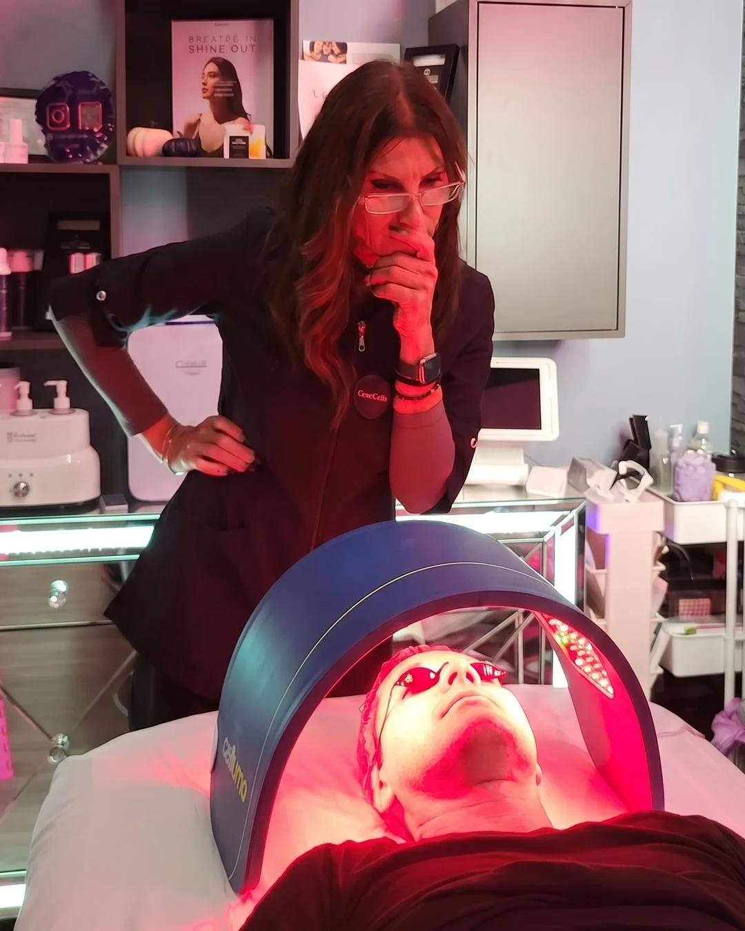 A woman with glasses and long hair, wearing a black jacket, is observing a man lying on a bed under a red light therapy device in a medical or spa setting. The man is wearing protective eyewear and lying with his eyes closed, while the woman appears to be concerned or thoughtful.