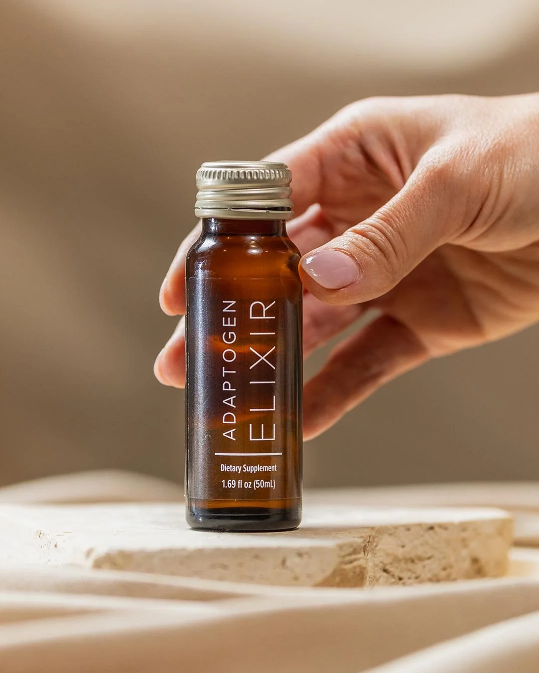 A person holding a small brown glass bottle labeled 'ADAPTOGEN IELIXIR' with a silver cap, standing on a wooden surface.