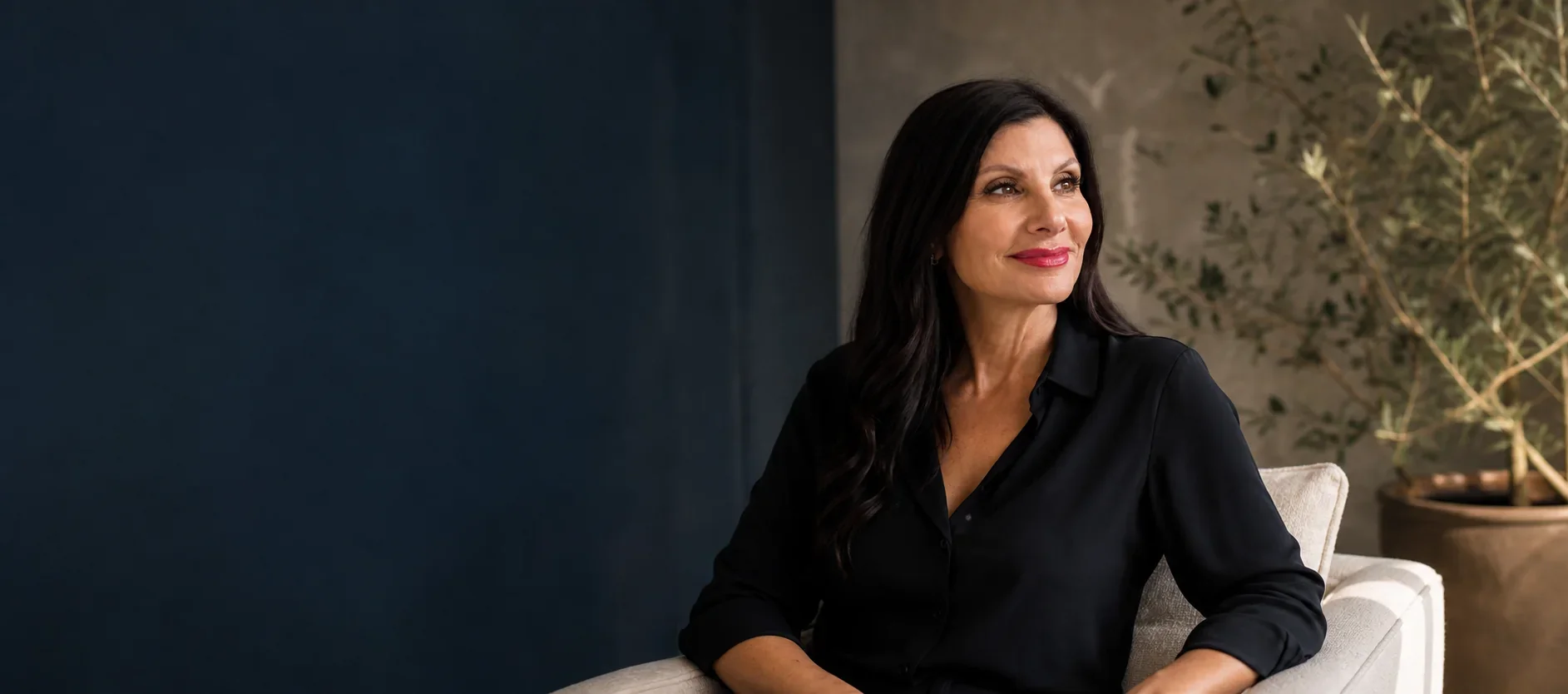A woman with dark hair and red lipstick sitting on a beige armchair, looking to her right with a pleasant expression, wearing a black shirt, with potted plant and textured wall in the background.