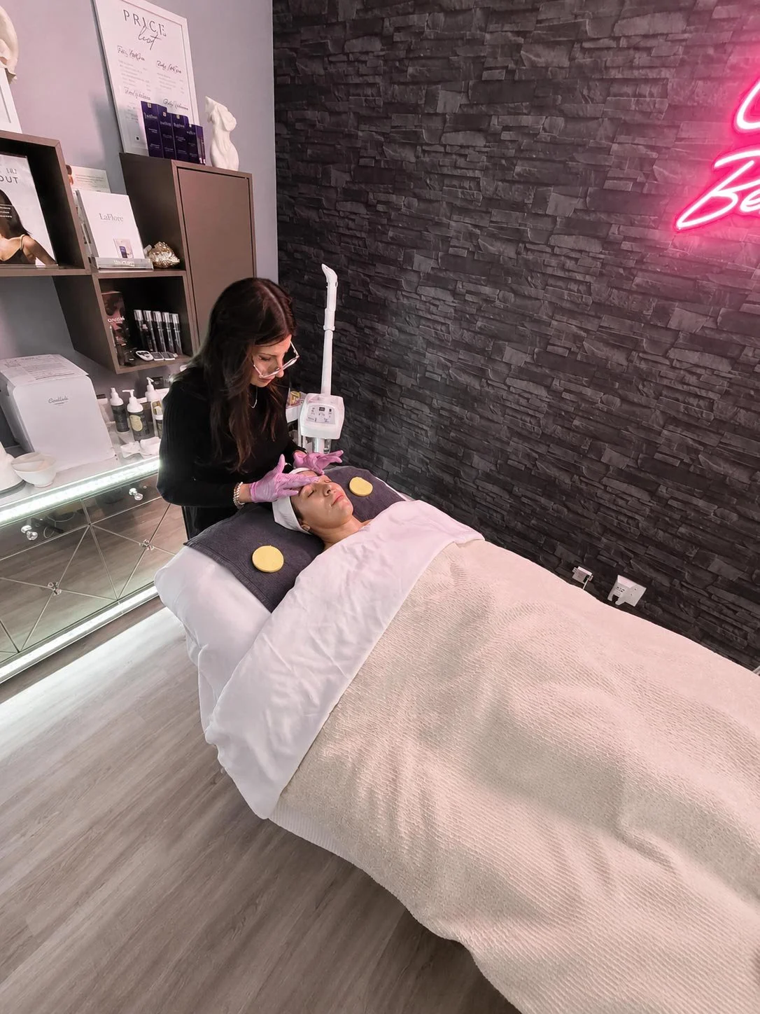 A woman lying on a treatment bed receiving a facial treatment from an esthetician in a spa or skincare clinic. The esthetician is wearing pink gloves and a black outfit. The treatment room has a black stone wall, shelves with skincare products, and a pink neon sign.