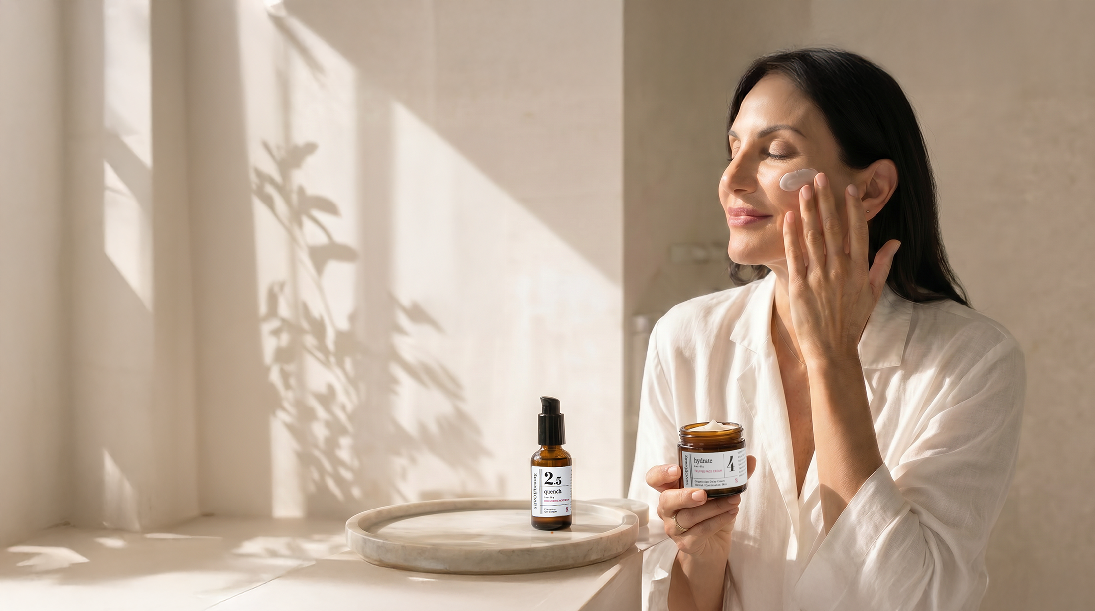 A woman with dark hair applying skincare cream to her face in a bright, minimalist bathroom with soft shadows and natural light.