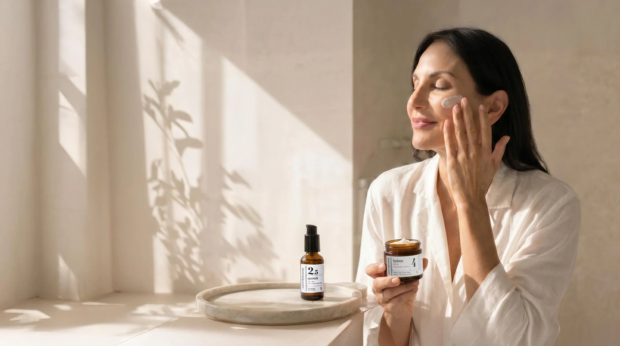 A woman in a white shirt applies cream to her face using her fingers in a softly lit room with beige and white tones.