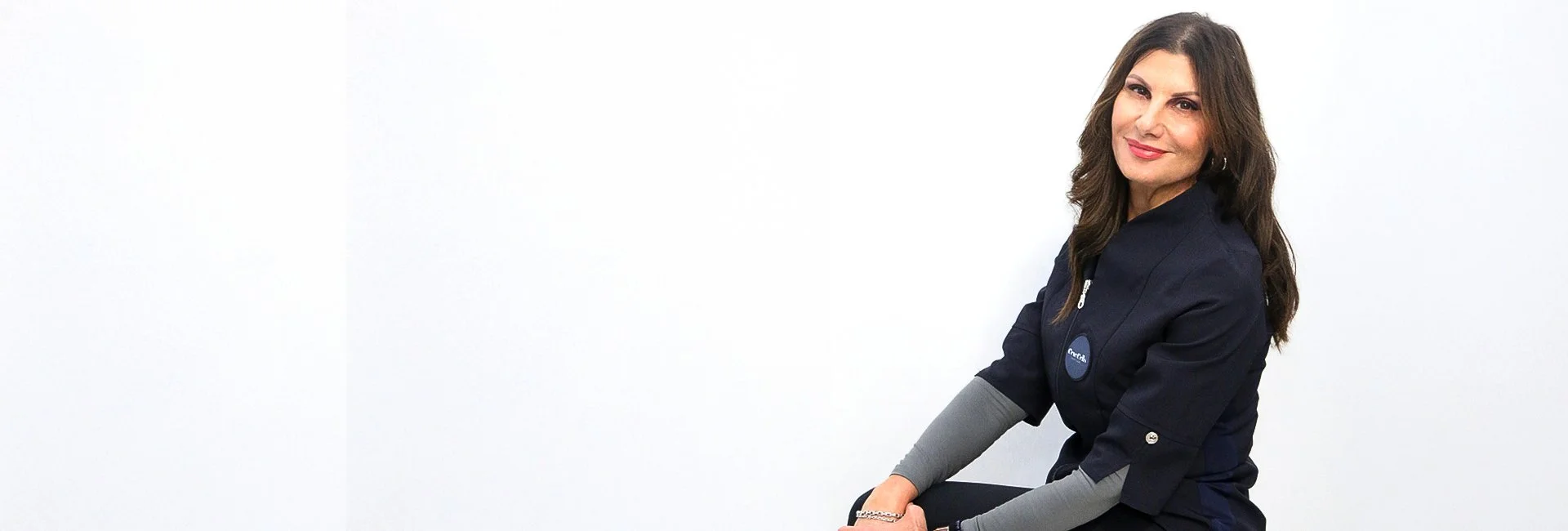 A woman with long brown hair, wearing a navy blue jacket and gray long sleeves, sitting against a white wall and smiling at the camera.