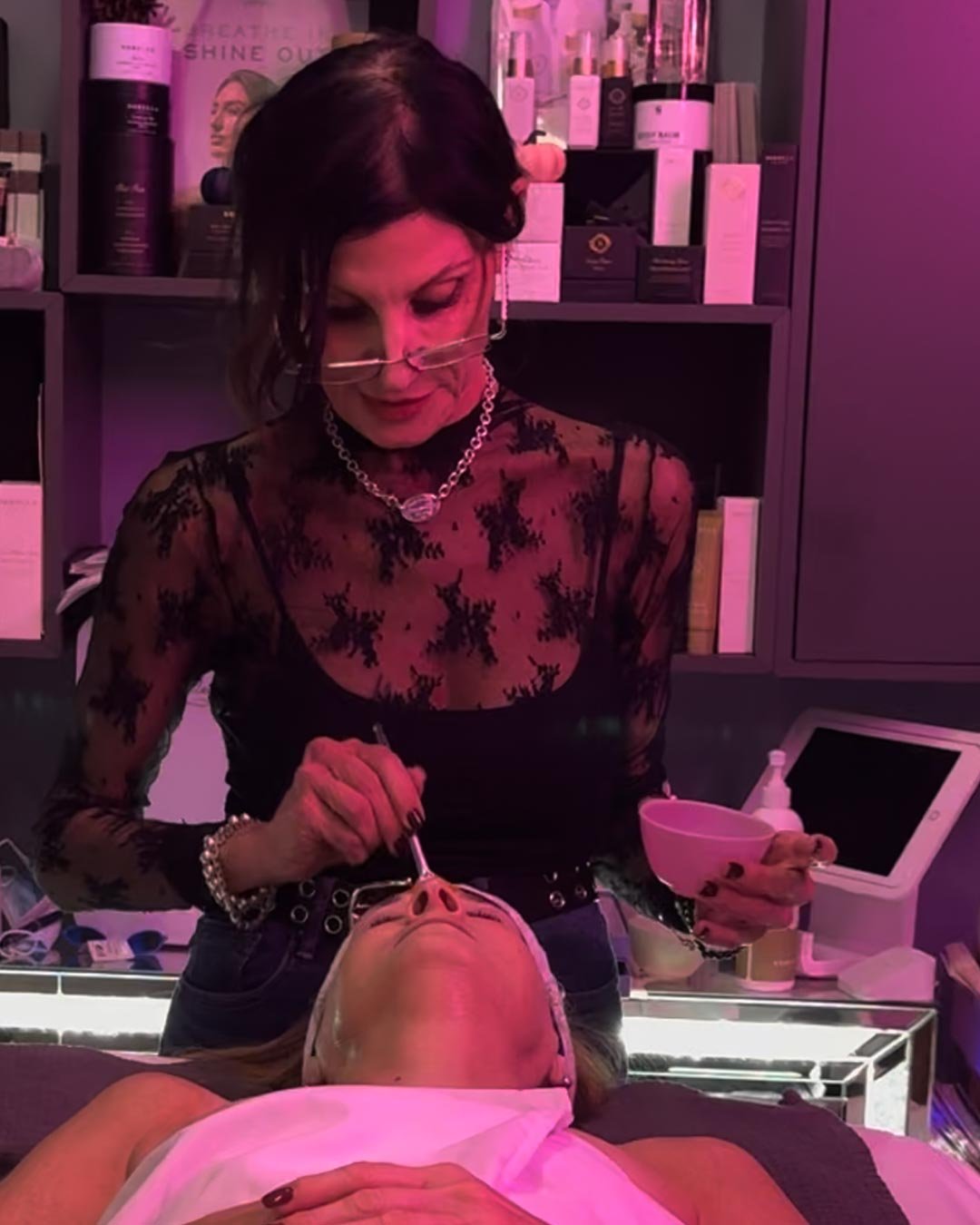 A woman in black lace top and jewelry provides a facial treatment to a patient lying down with eyes closed in a spa or skincare clinic, with shelves of skincare products in the background.