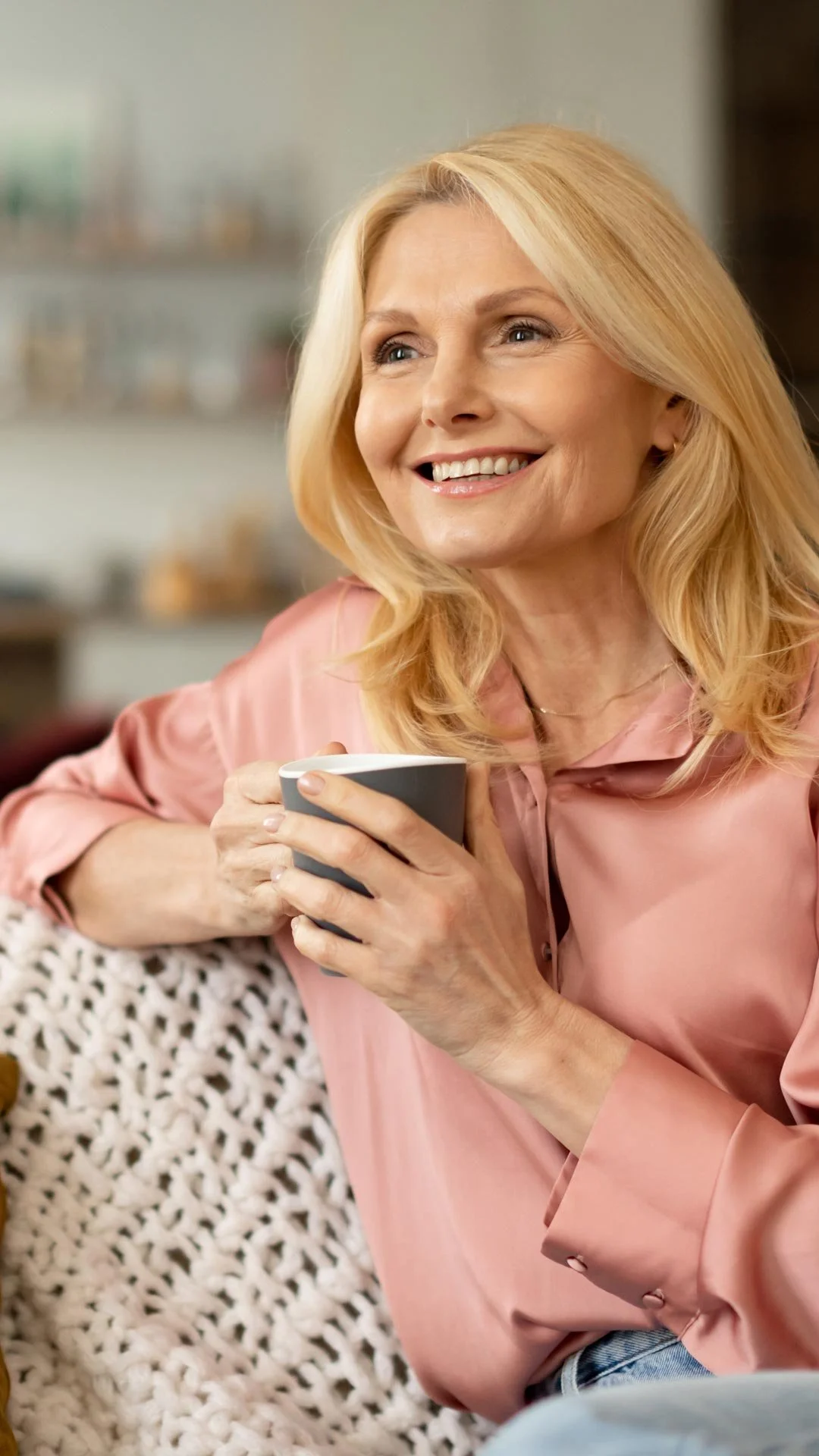An older woman with blonde hair and a pink satin blouse smiling while holding a gray coffee mug in a cozy room.