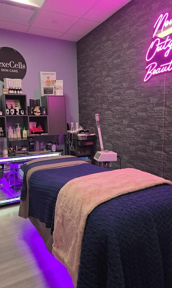 A beauty treatment room with a massage bed covered with a navy blue quilt and a beige blanket, illuminated by purple lighting. The wall behind has a neon sign and a textured gray wall. Shelves with skincare products and equipment are visible, indicating a skincare or beauty clinic environment.