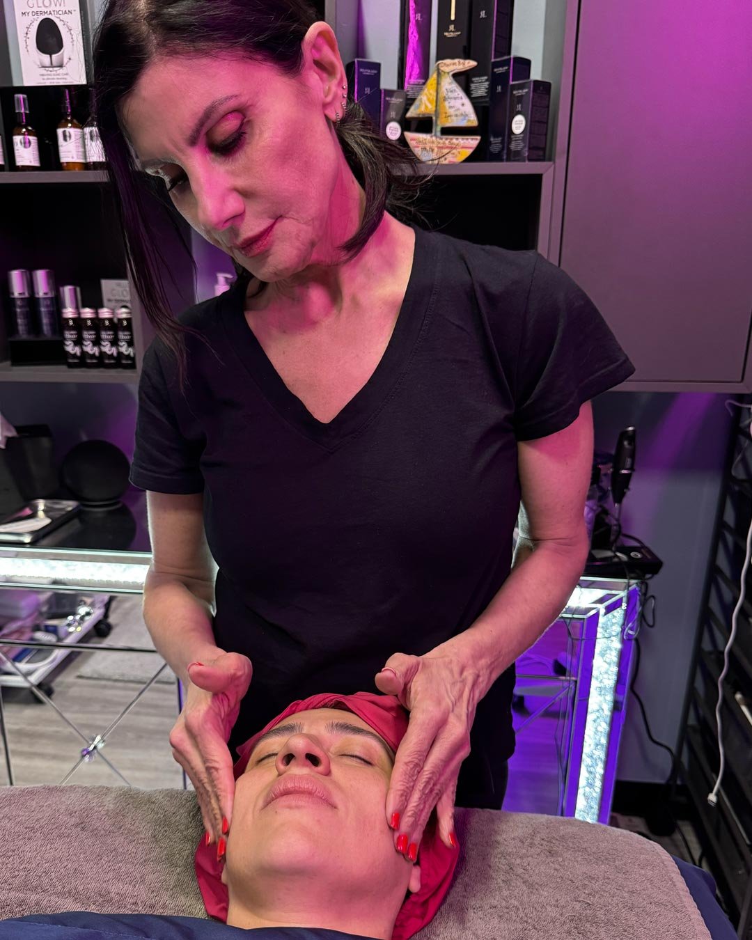 A woman receiving a facial massage at a spa or skincare clinic, with shelves of skincare products in the background.