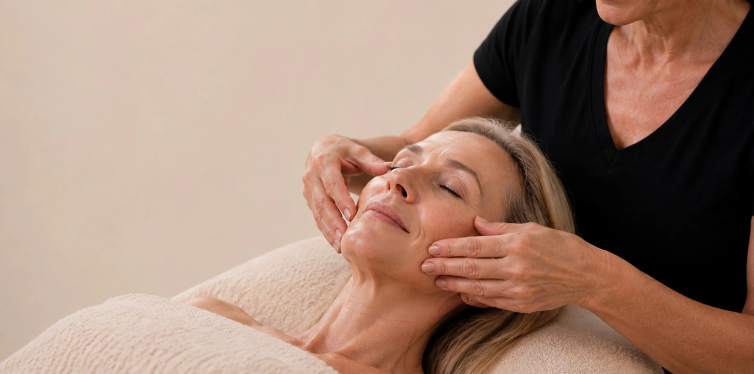 A woman receives a facial treatment from a cosmetologist, lying on her back with her eyes closed, while the cosmetologist gently massages her face in a room with beige walls.