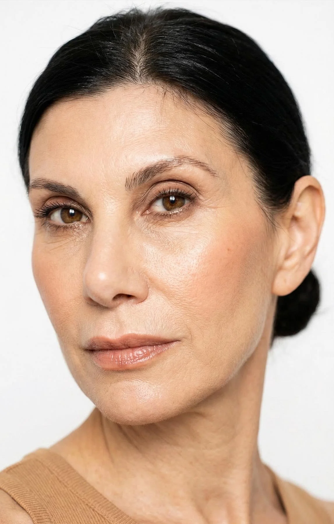 Close-up of a woman with fair skin, dark hair styled back, brown eyes, natural makeup, and a beige top, against a plain white background.