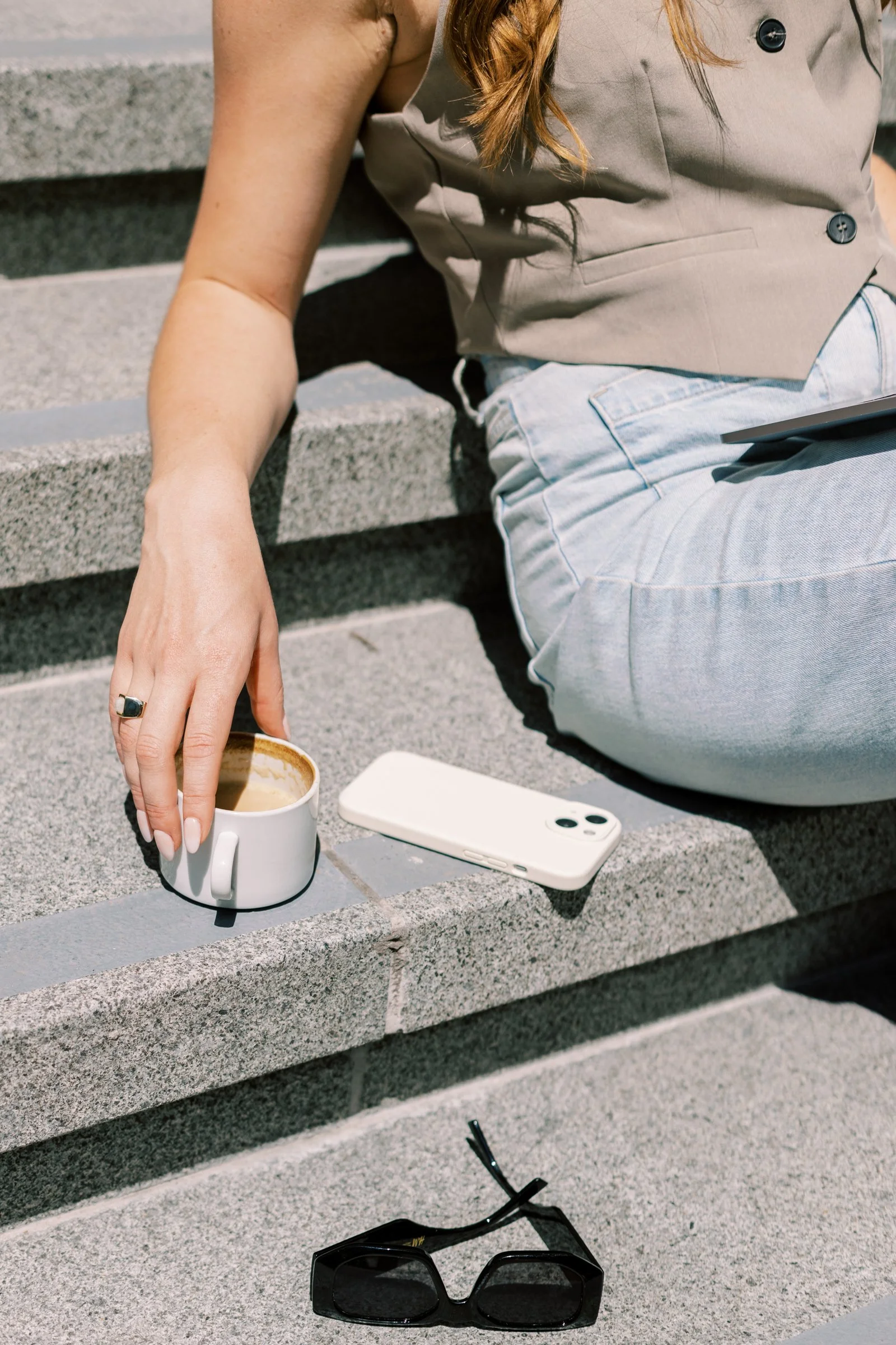 A person sitting on outdoor concrete stairs, wearing a beige sleeveless vest and light blue jeans. They are holding a coffee mug and have a white smartphone and sunglasses on the stairs.