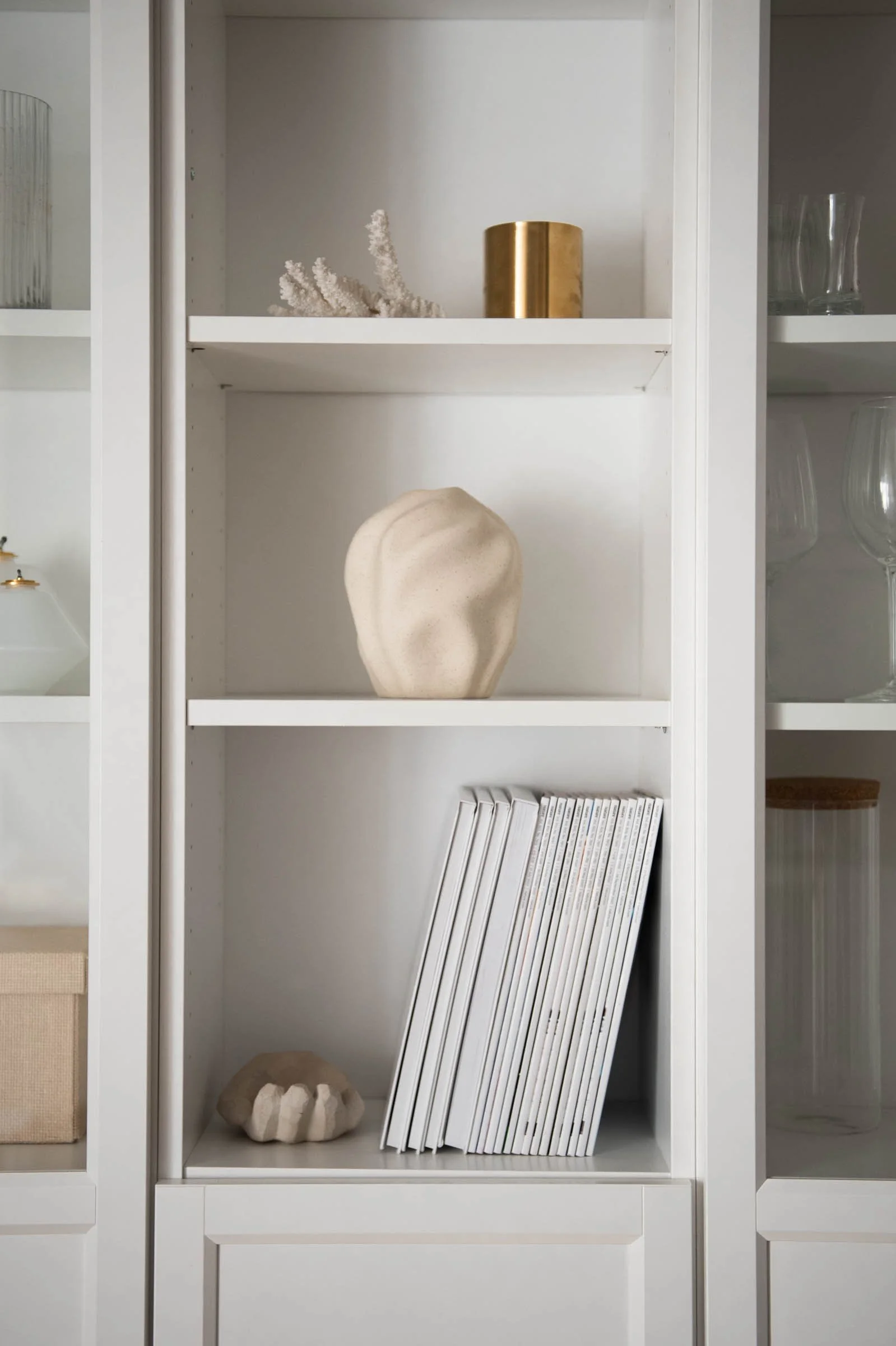 White cabinet with decorative items including a gold candle holder, white coral, a beige stone sculpture, and a row of white books or magazines.
