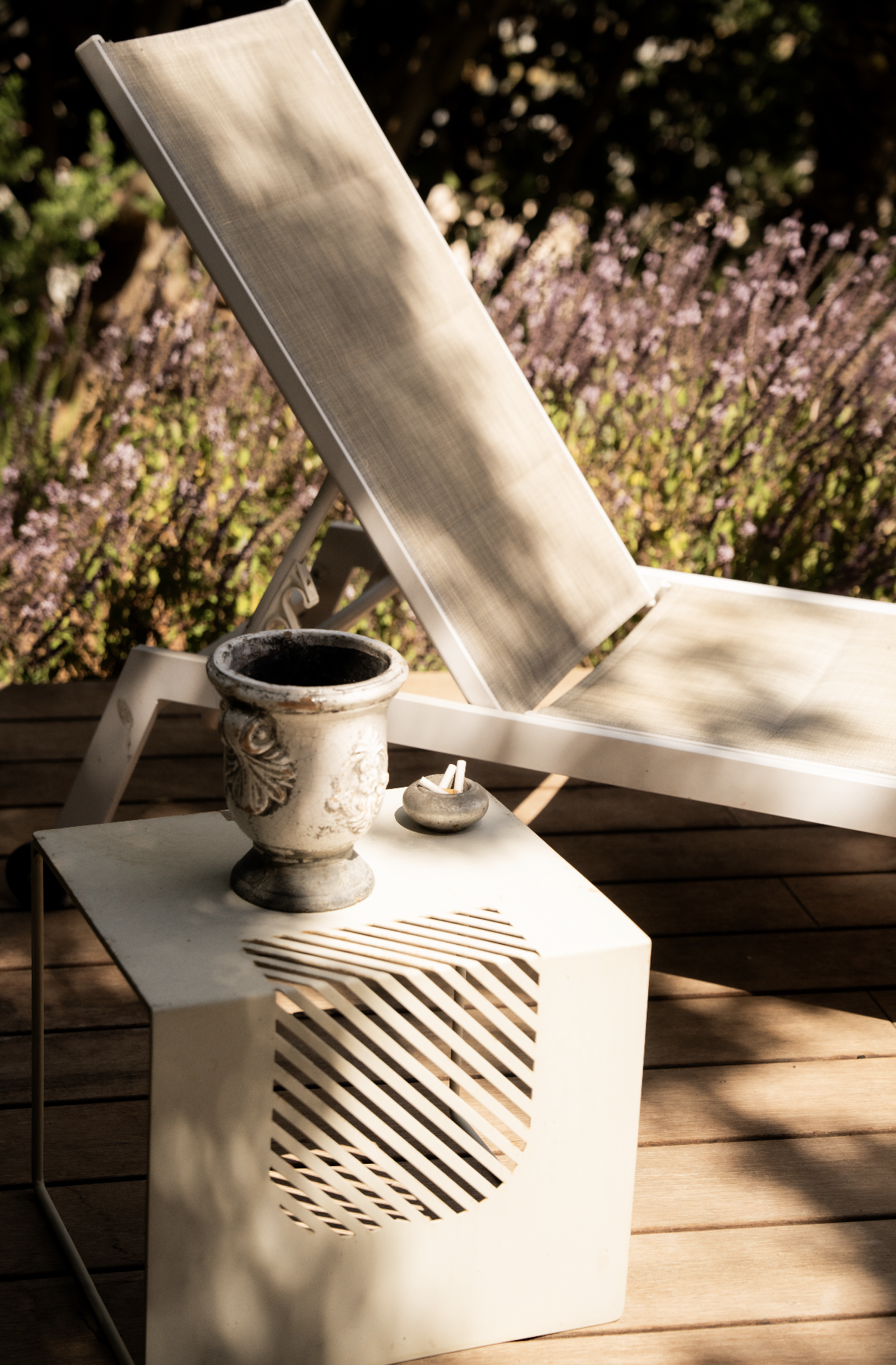 A white lounge chair on a wooden deck, with a small table holding a decorative urn, a small dish, and a comb, outdoor background with purple flowers and dark tree shadows.