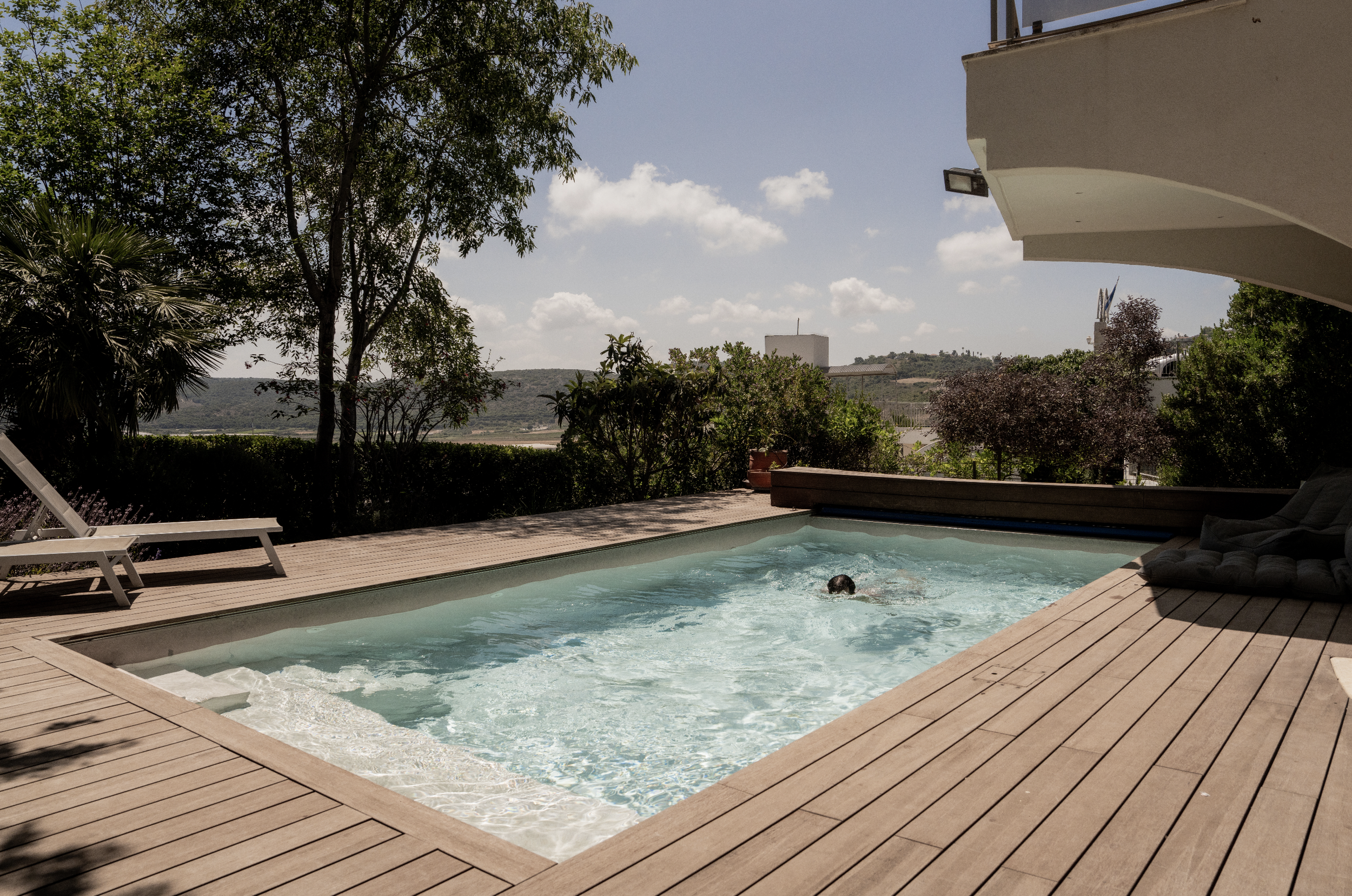 A backyard with a swimming pool and a wooden deck, surrounded by trees and hills, under a partly cloudy sky.