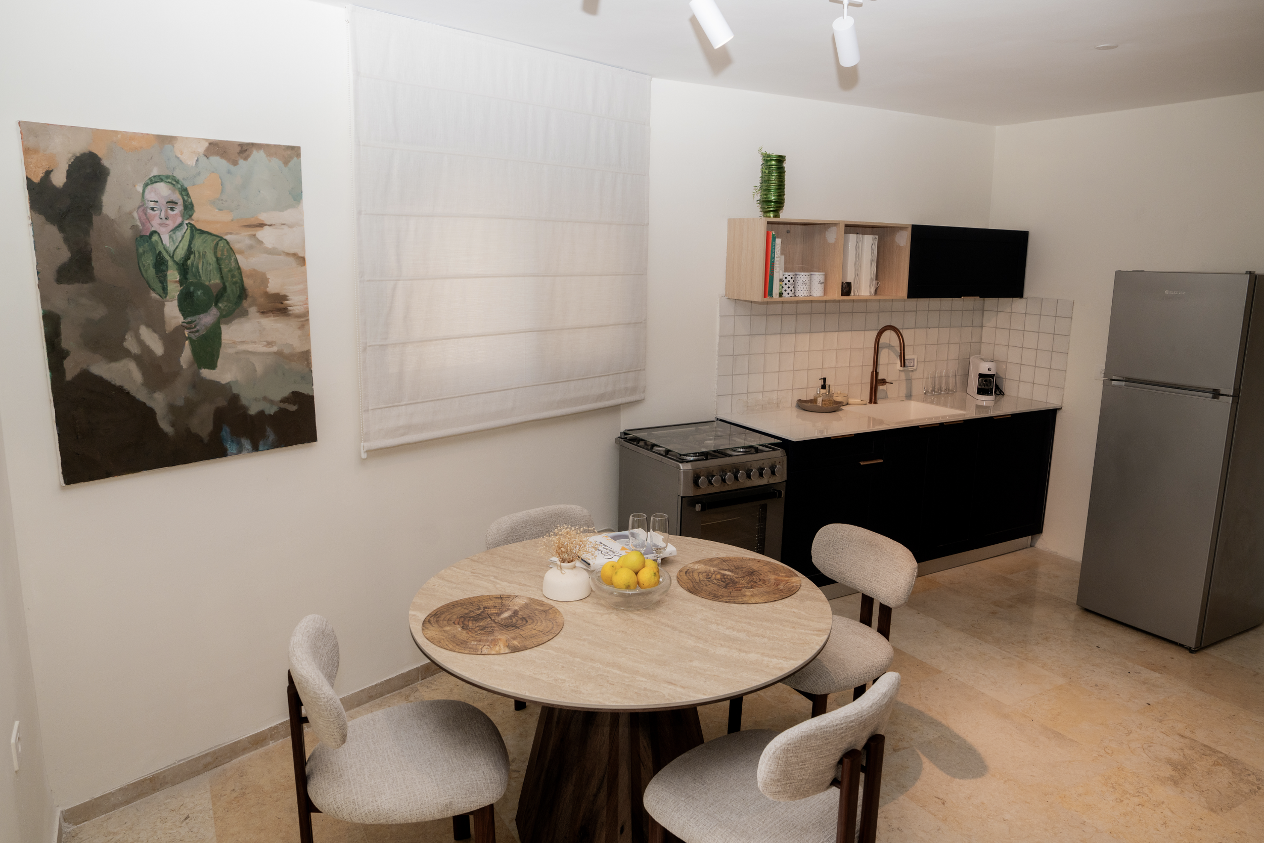 A round dining table with six upholstered chairs, a bowl of lemons, and glasses, set in a kitchen area with a stove, refrigerator, and wall-mounted shelf with books and decorative items.