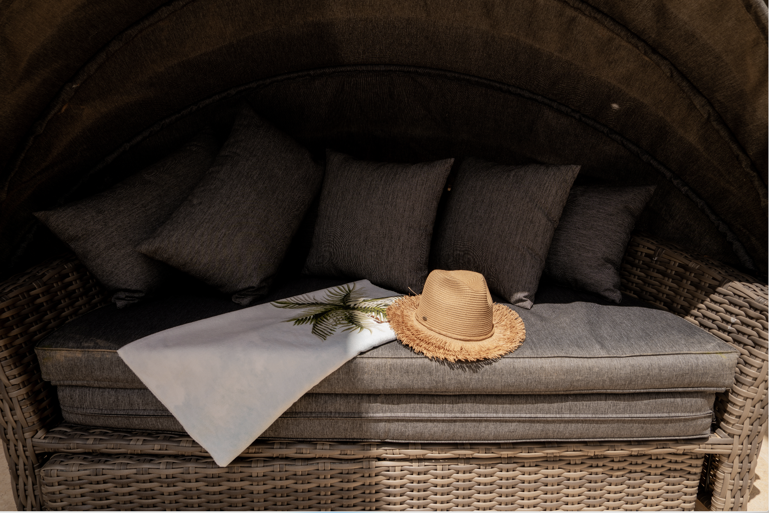 A wicker outdoor sofa with dark cushions, a straw hat, a white towel with green leaves, and a white cloth on it, situated under a canopy with dark fabric.