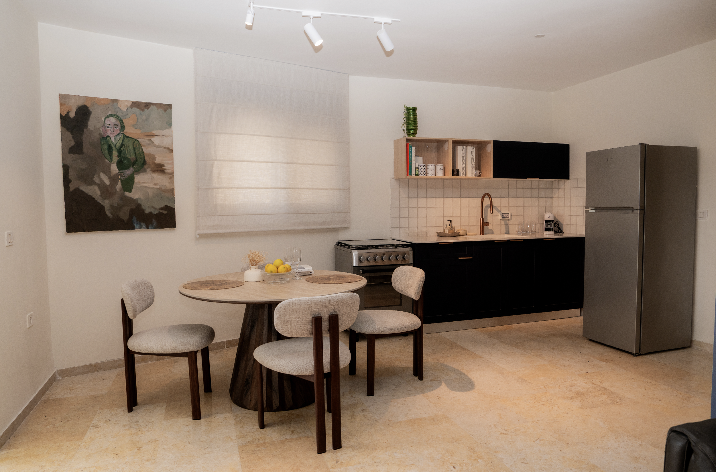 A modern kitchen and dining area with a round wooden table surrounded by four beige upholstered chairs. On the table is a glass bowl with lemons, a small potted plant, and two glasses. The kitchen has black lower cabinets, an open wooden shelf with b