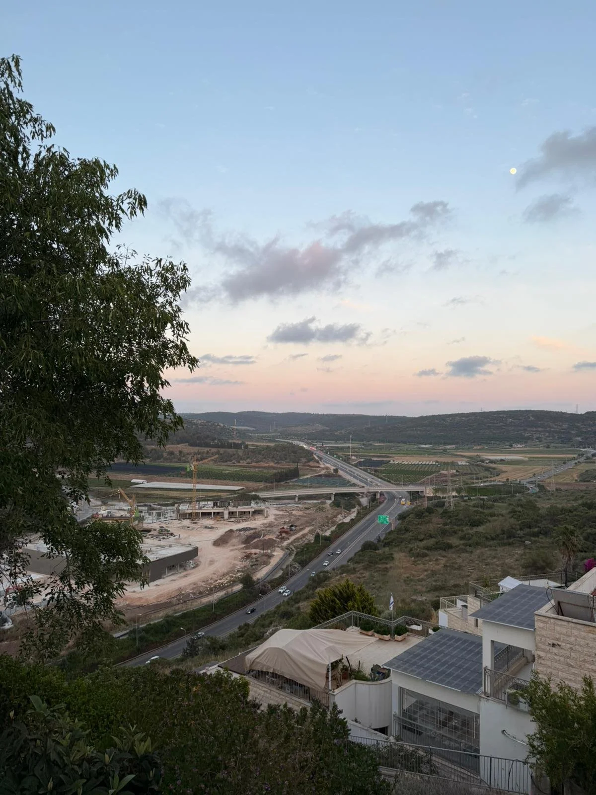 View of a landscape with hills, a highway, and construction site during sunset with a partly cloudy sky and visible moon.