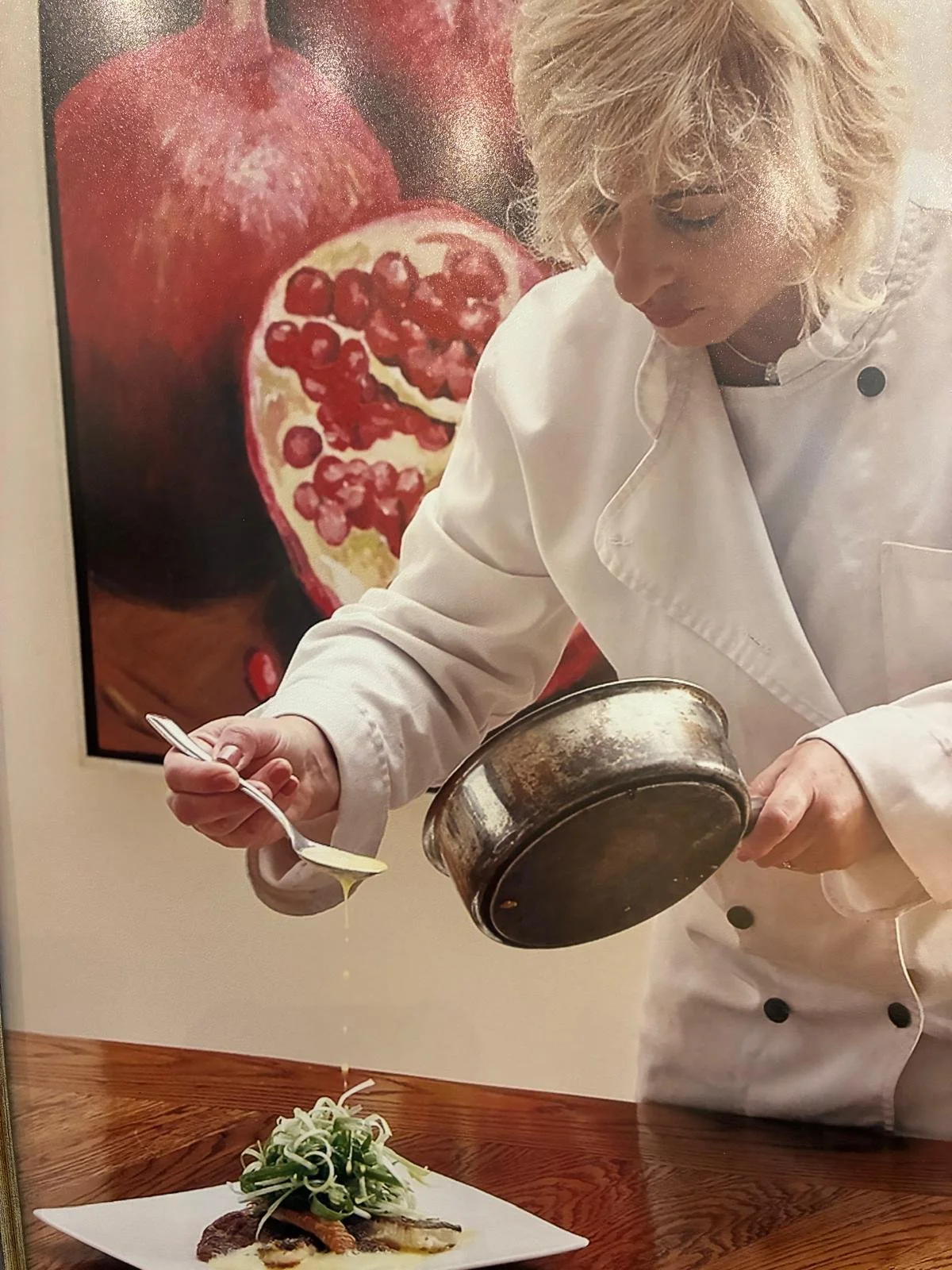 Chef in white coat pouring sauce over a plated dish in a kitchen with pomegranate artwork in the background.
