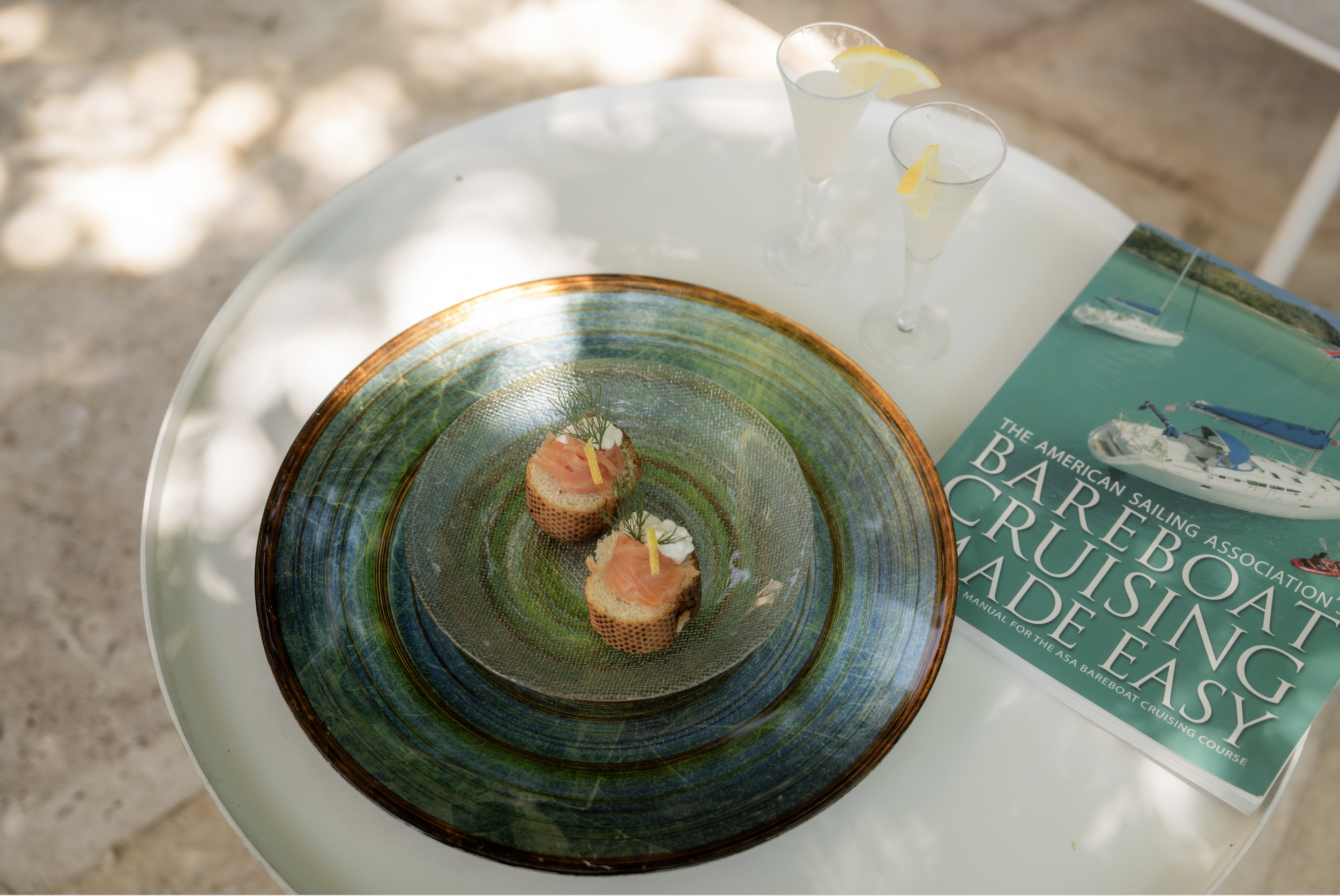 Three small appetizers with smoked salmon, cream cheese, and dill on toasted bread sitting on a colorful glass plate on a white table. Two lemon drinks in tall glasses with lemon slices and a boat cruise guidebook titled 'The American Sailing Associa