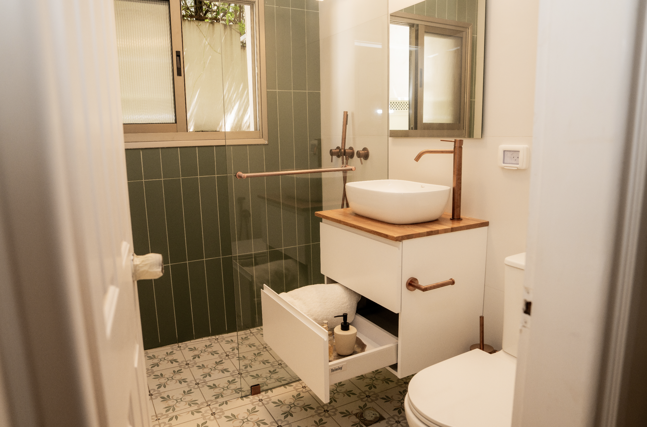 A small bathroom with green tiled walls, a white wash basin on a wooden countertop, a mirror, a window, and patterned floor tiles.