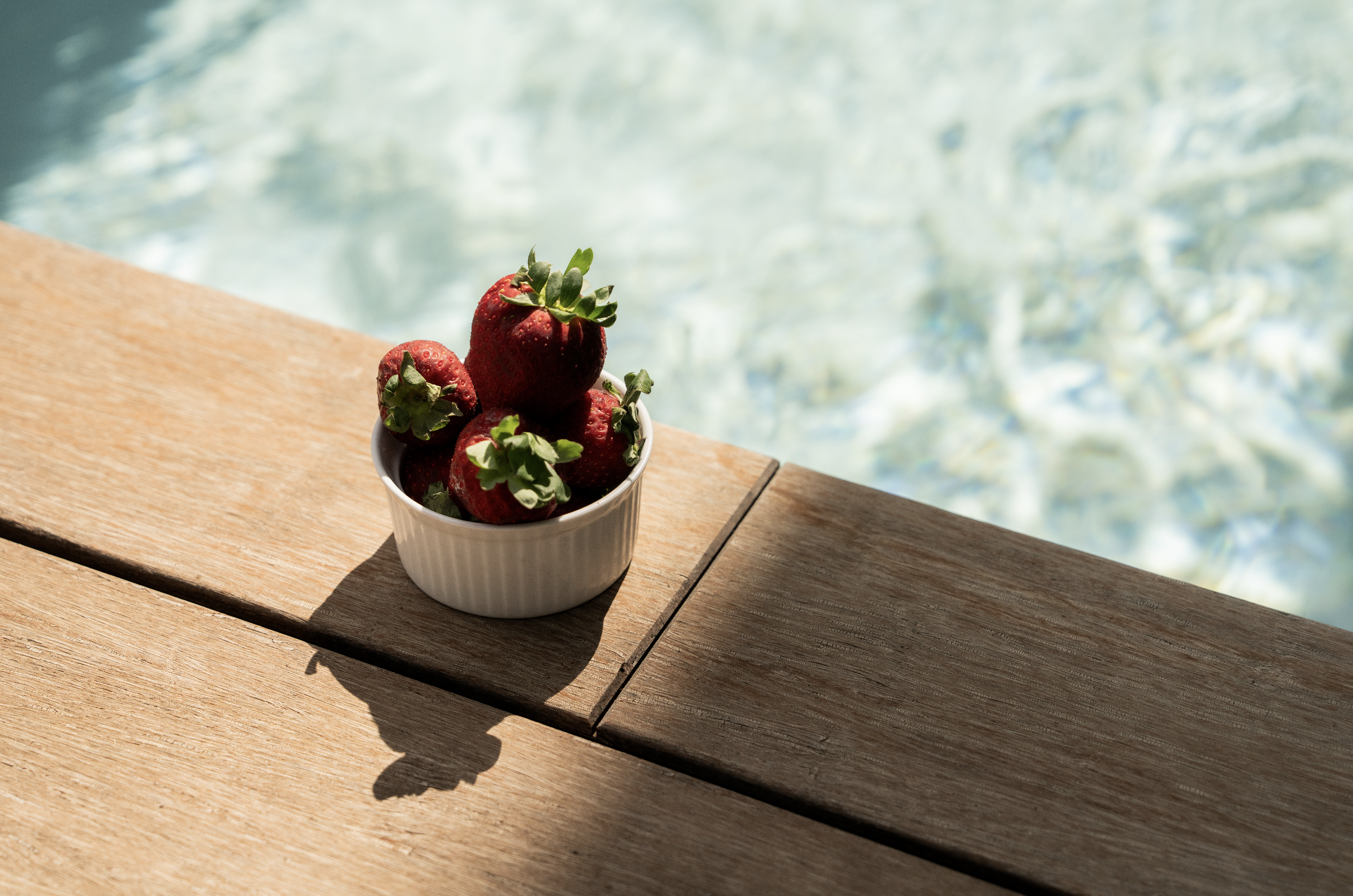 A small white bowl filled with fresh strawberries on a wooden surface, with sunlight casting shadows and a blurry blue and white background