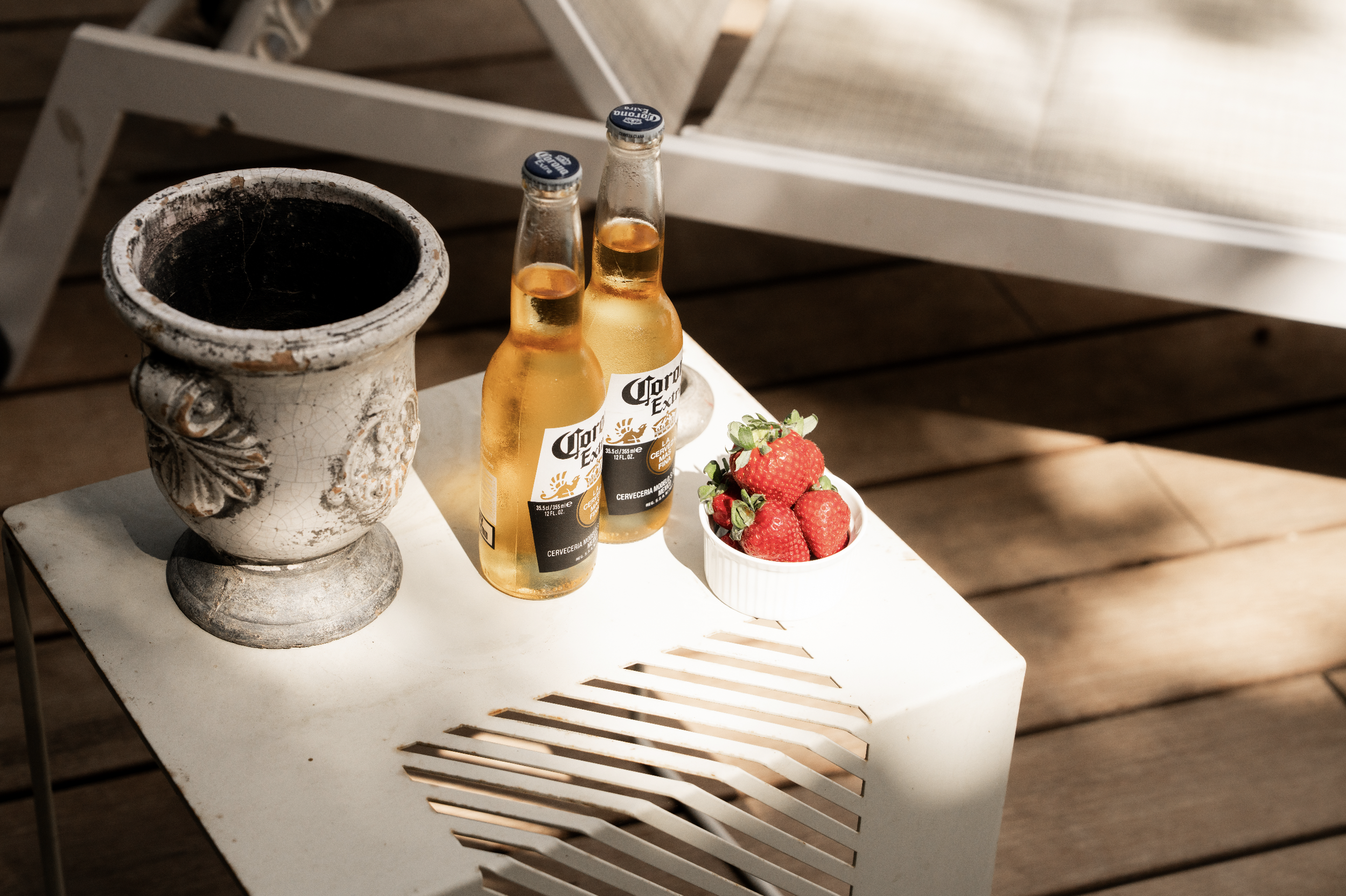 A white outdoor table holding two bottles of Corona beer, a bowl of strawberries, and an antique ceramic mug, with a wooden deck and outdoor furniture in the background.