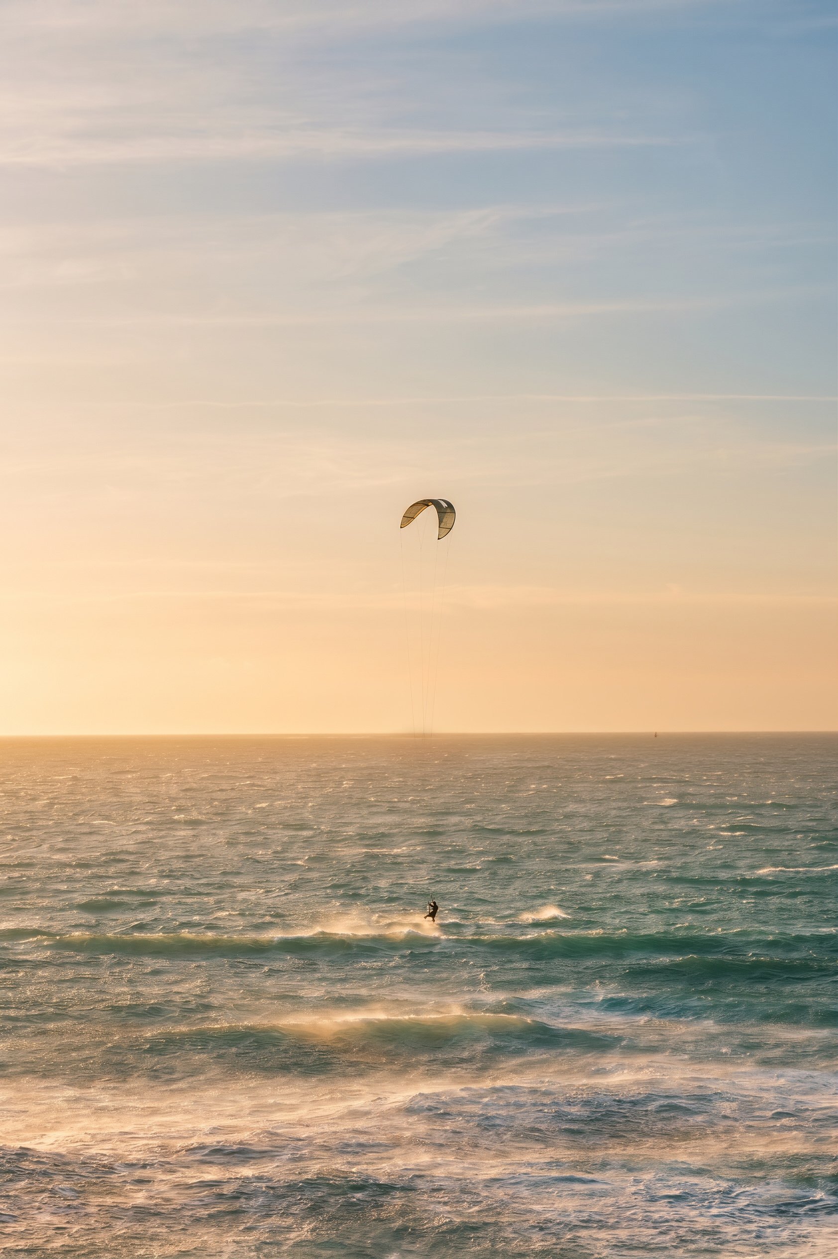 A person kite surfing on the ocean during sunset, with a kite in the sky and waves below.