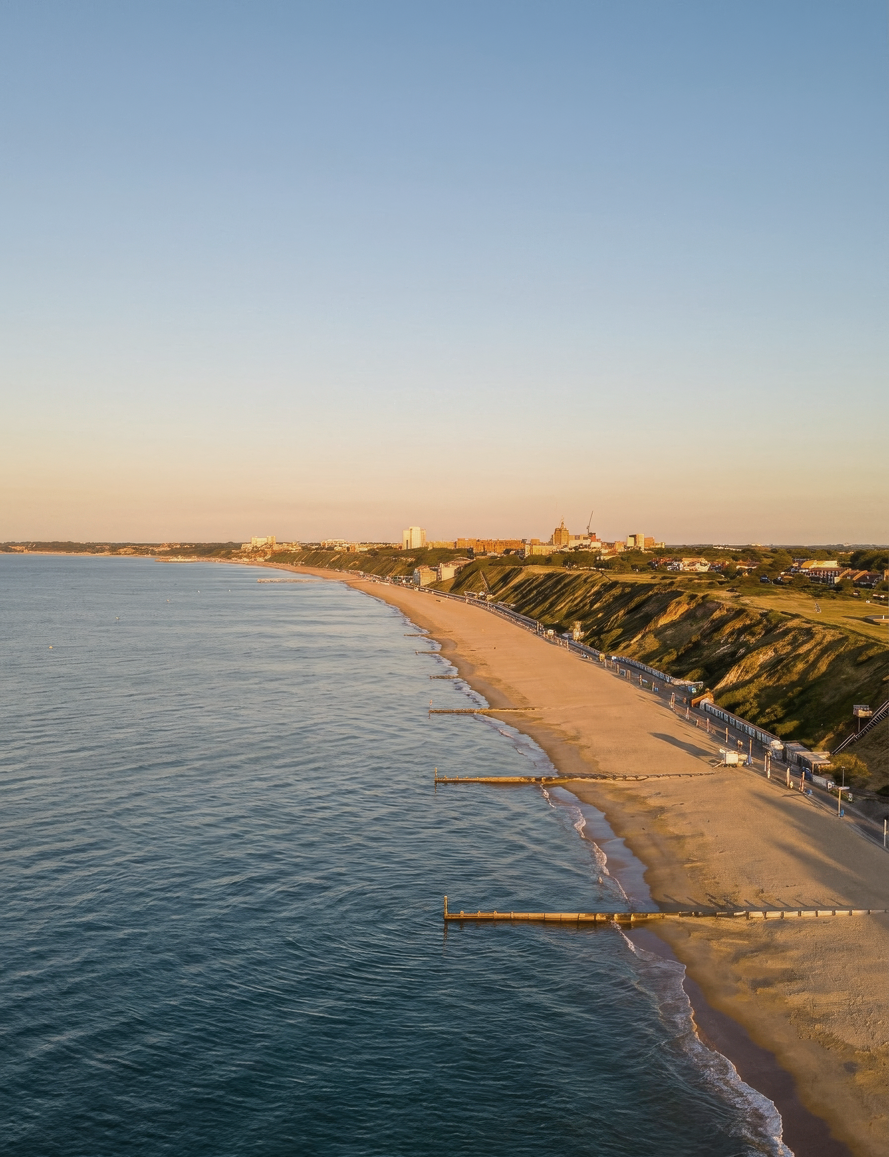 A coastline with a sandy beach, a promenade with barriers along it, and a cityscape with buildings and green hills in the background, under a clear sky.