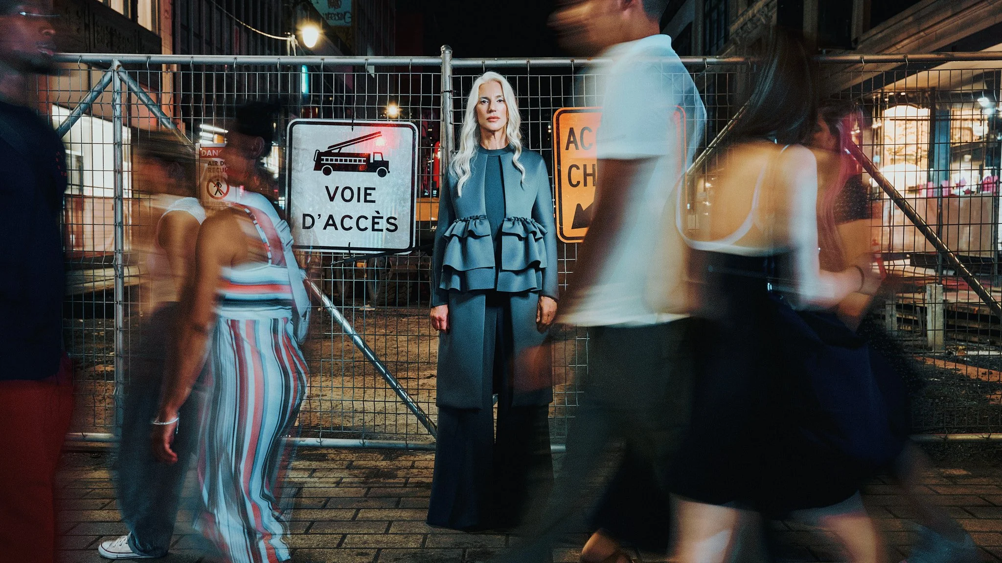 Model standing sharp and still in Maison Marie Saint Pierre while pedestrians blur past a construction barrier, Montreal fashion photography.