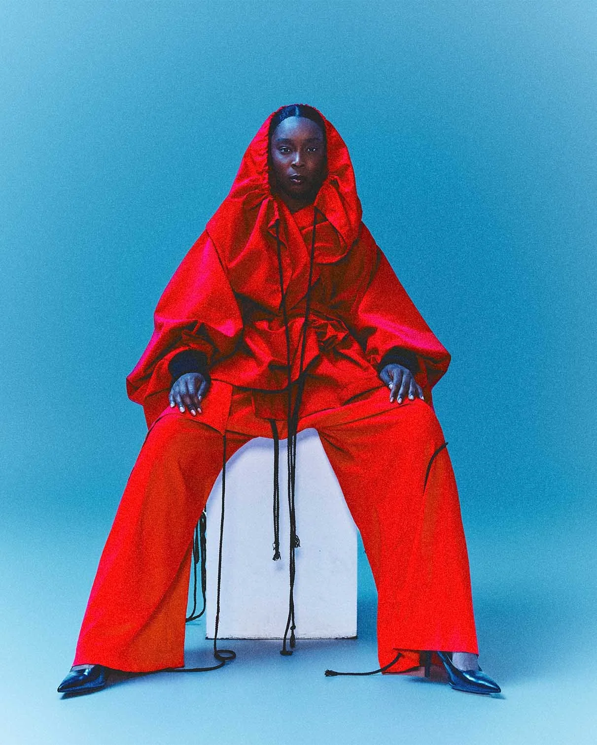 A person wearing a red oversized hooded jacket and matching wide-leg pants, sitting on a white block against a teal background.