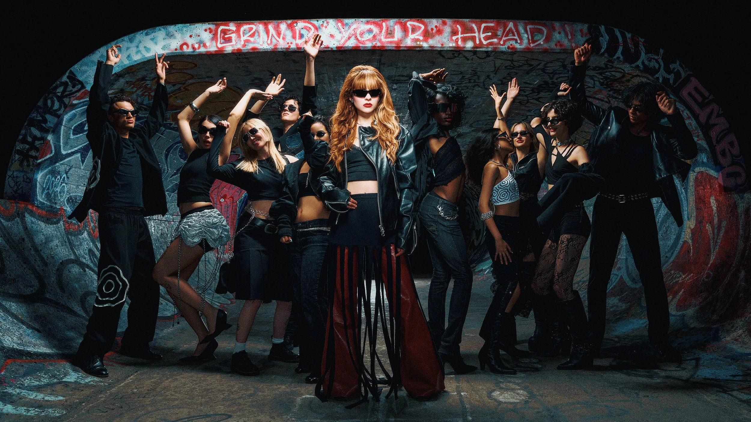 Group of stylish young women in black clothing and sunglasses posing in an urban alley with colorful graffiti, central woman with red hair and leather jacket, sign overhead reading 'GRIND YOUR HEAD'.