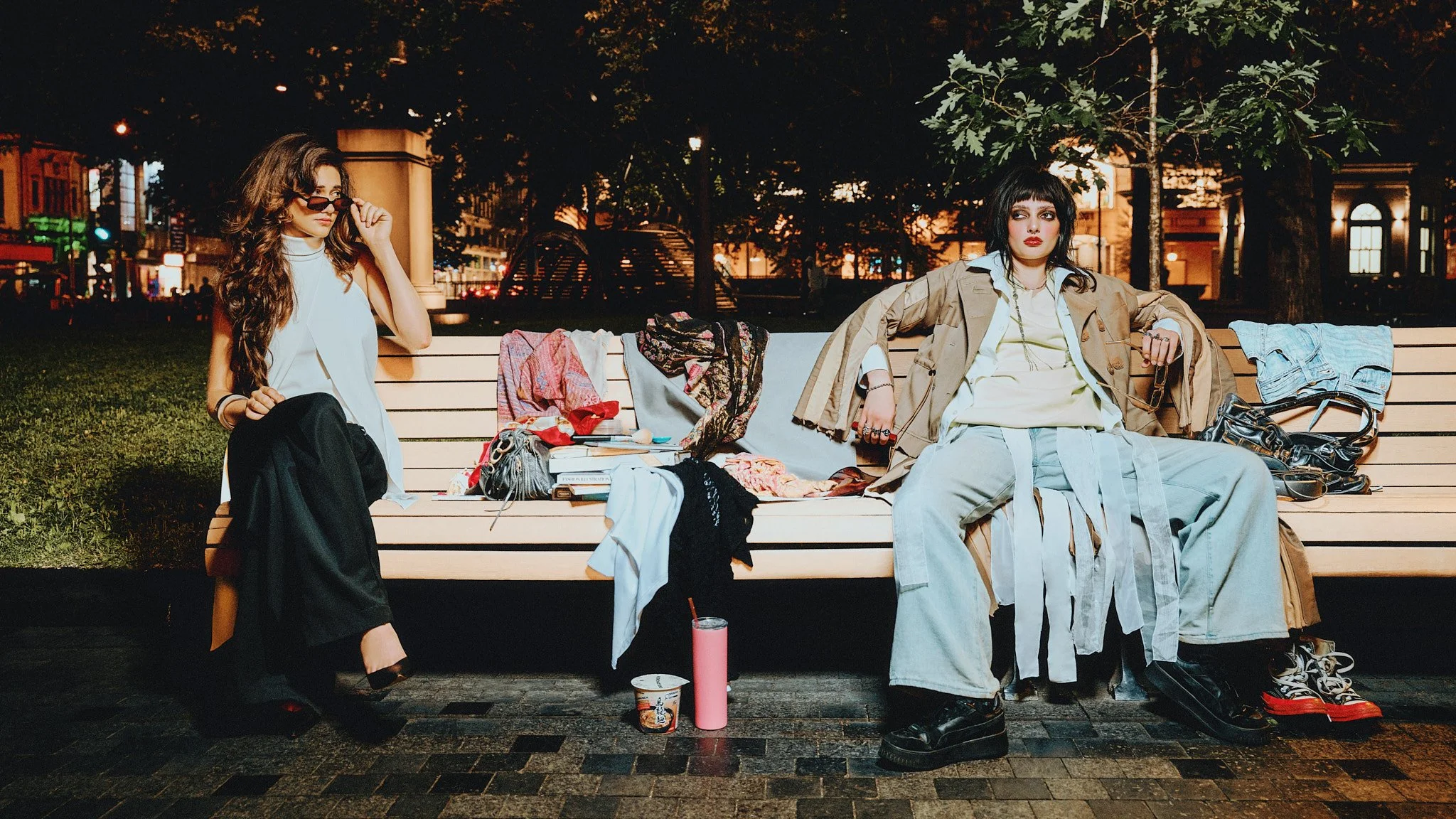 Two models seated at opposite ends of a park bench covered in personal belongings at night