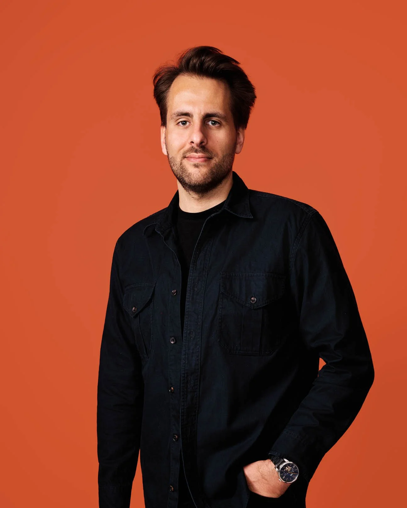A man with brown hair, beard, and light skin wearing a black jacket and watch, standing against an orange background.