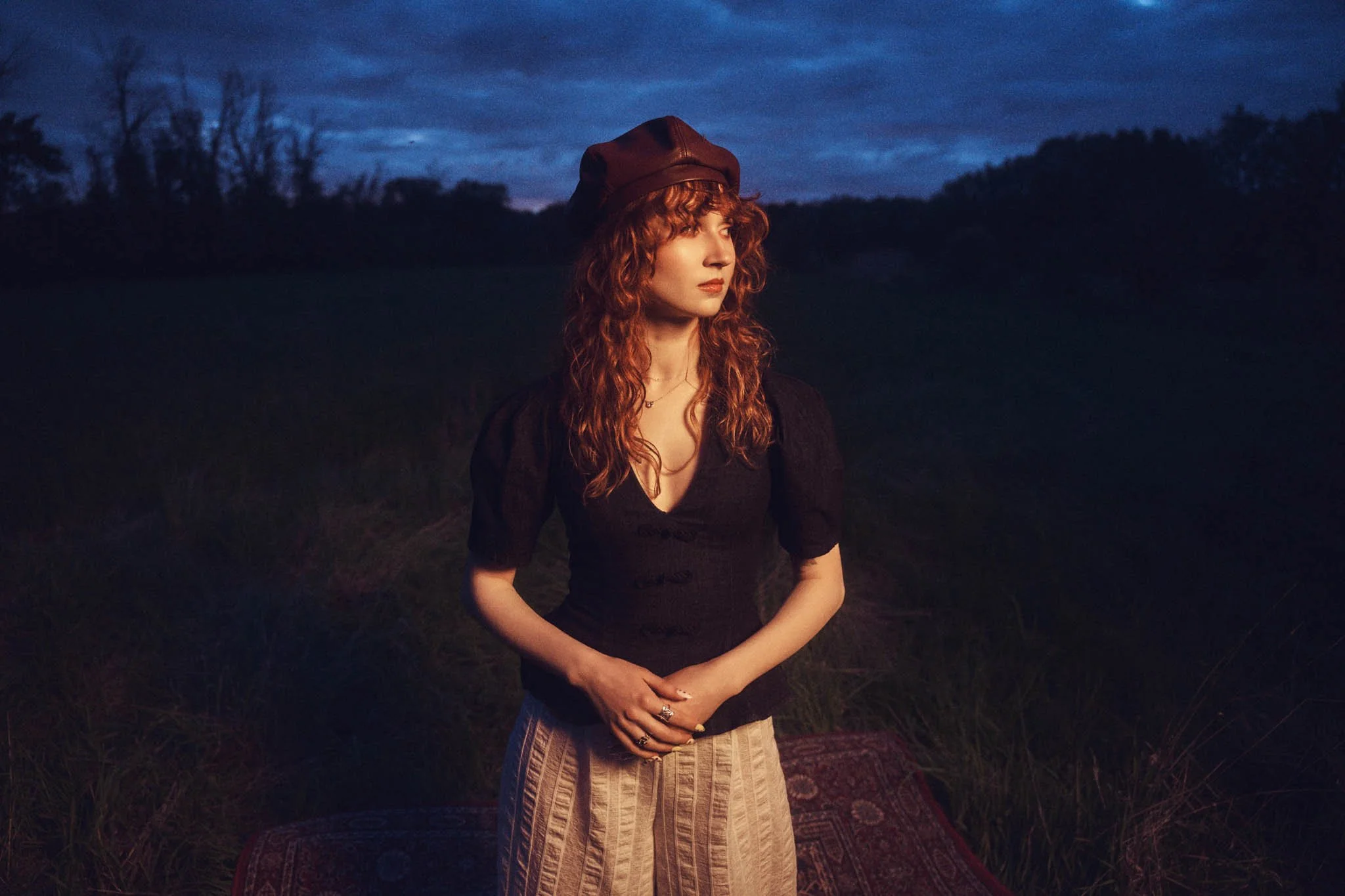 A woman with curly red hair wearing a dark top and a brown hat, standing outdoors at dusk with trees and a cloudy sky in the background.