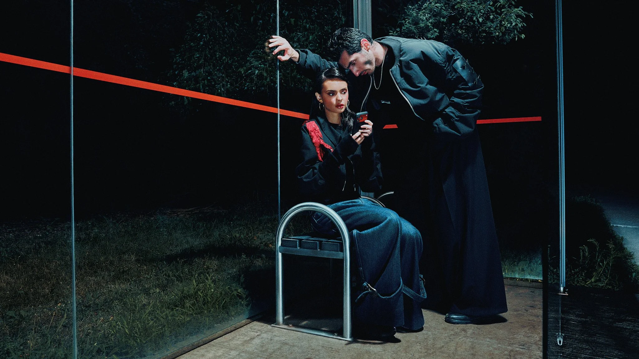 Model leaning over another looking at a phone screen inside a lit bus shelter at night