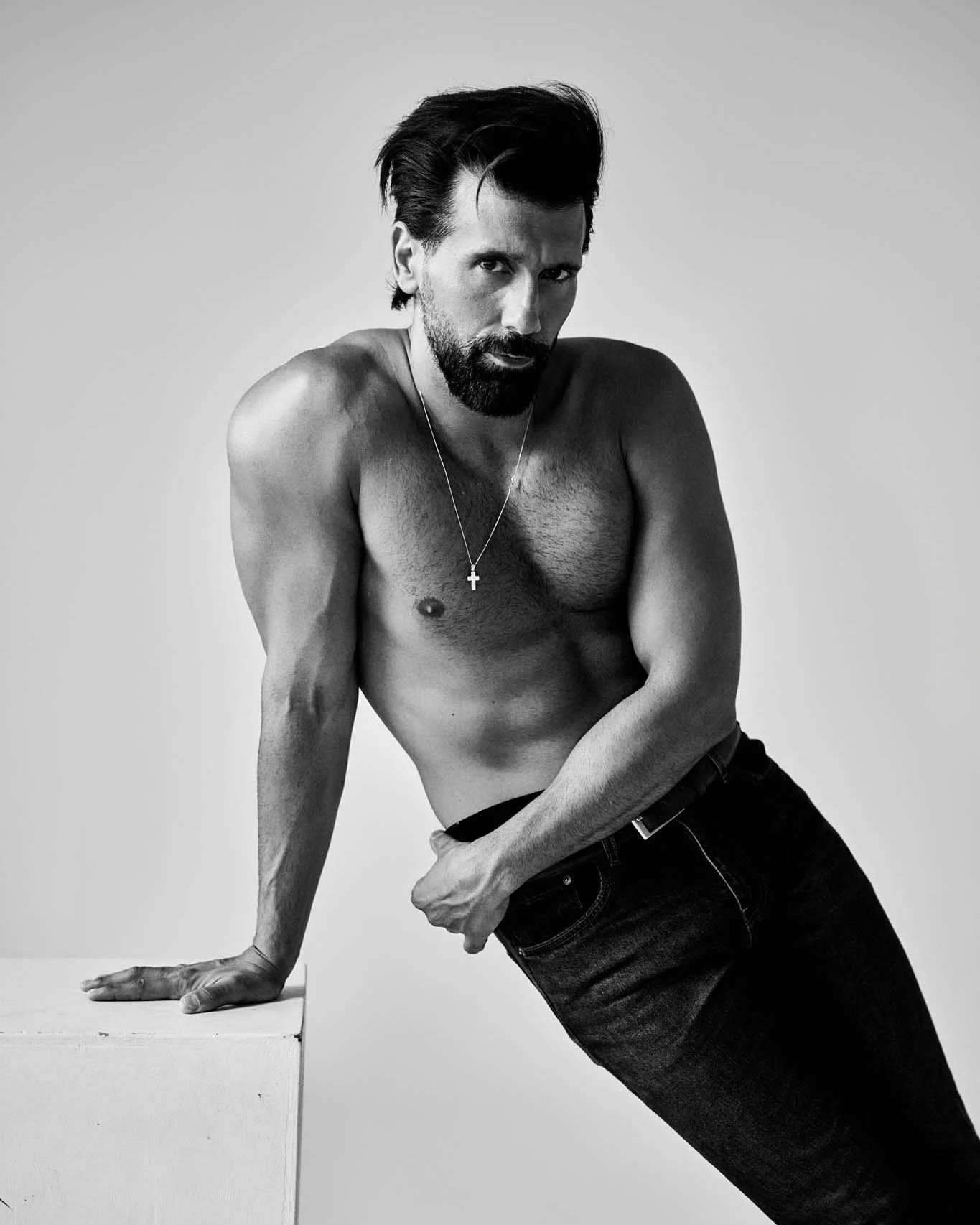 Black and white photo of a shirtless man with dark, styled hair and a beard, wearing a cross necklace. He is leaning on a white surface with his left arm, looking at the camera with an intense expression.