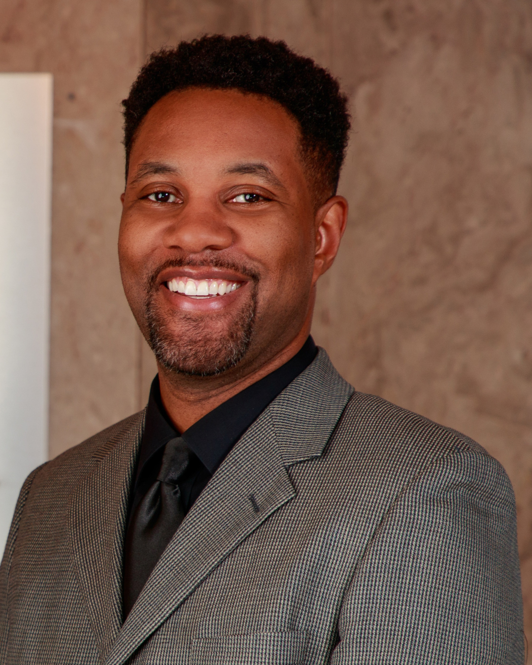 Chef Wayne G. Hooker Scholarship Board Member A smiling man dressed in a gray checkered suit and black shirt, posing against a neutral background.
