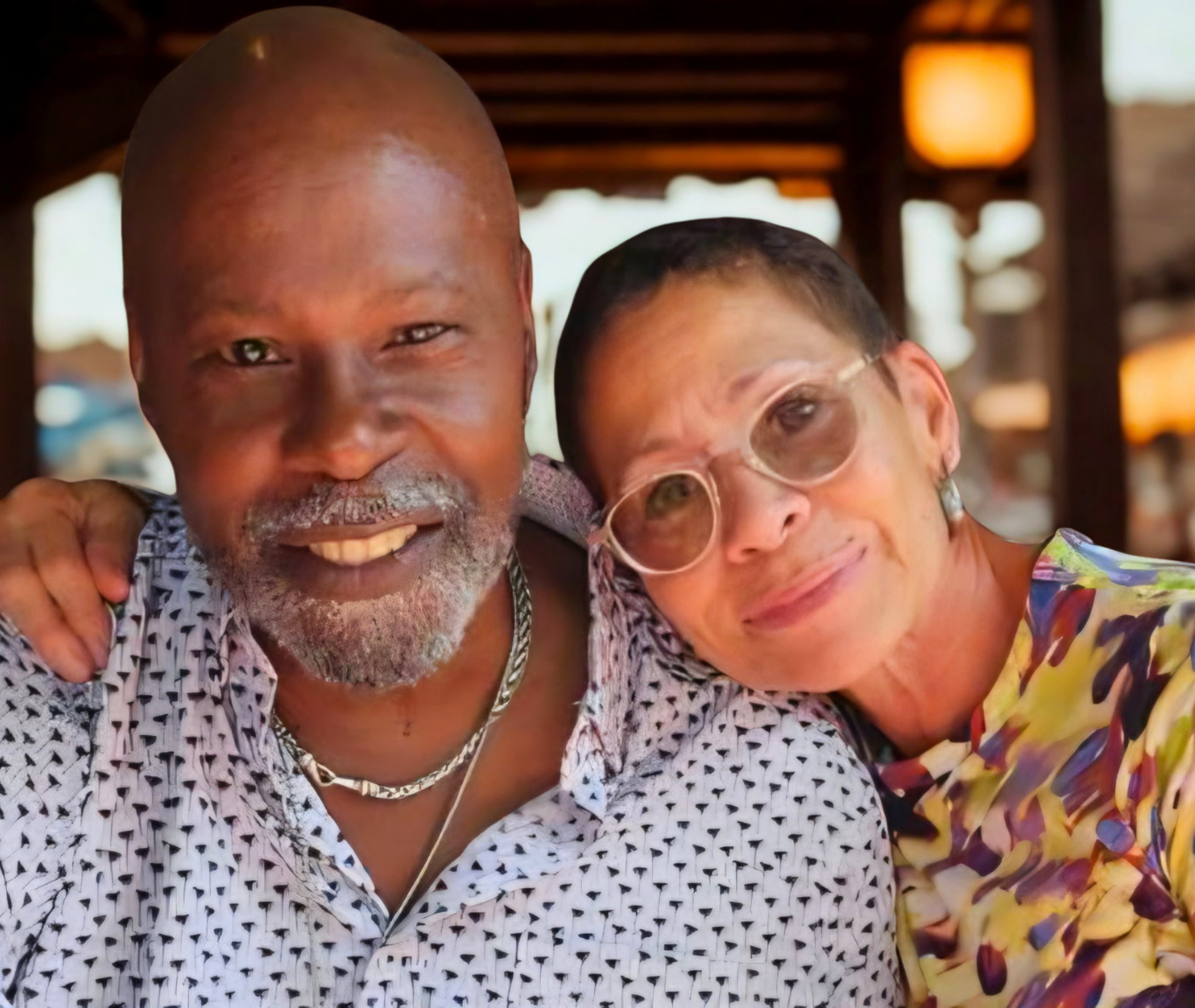Chef Wayne Hooker and founder Lisa Wyatt-Hooker, one wearing glasses, embrace each other in an outdoor setting with blurred warm lighting in the background.