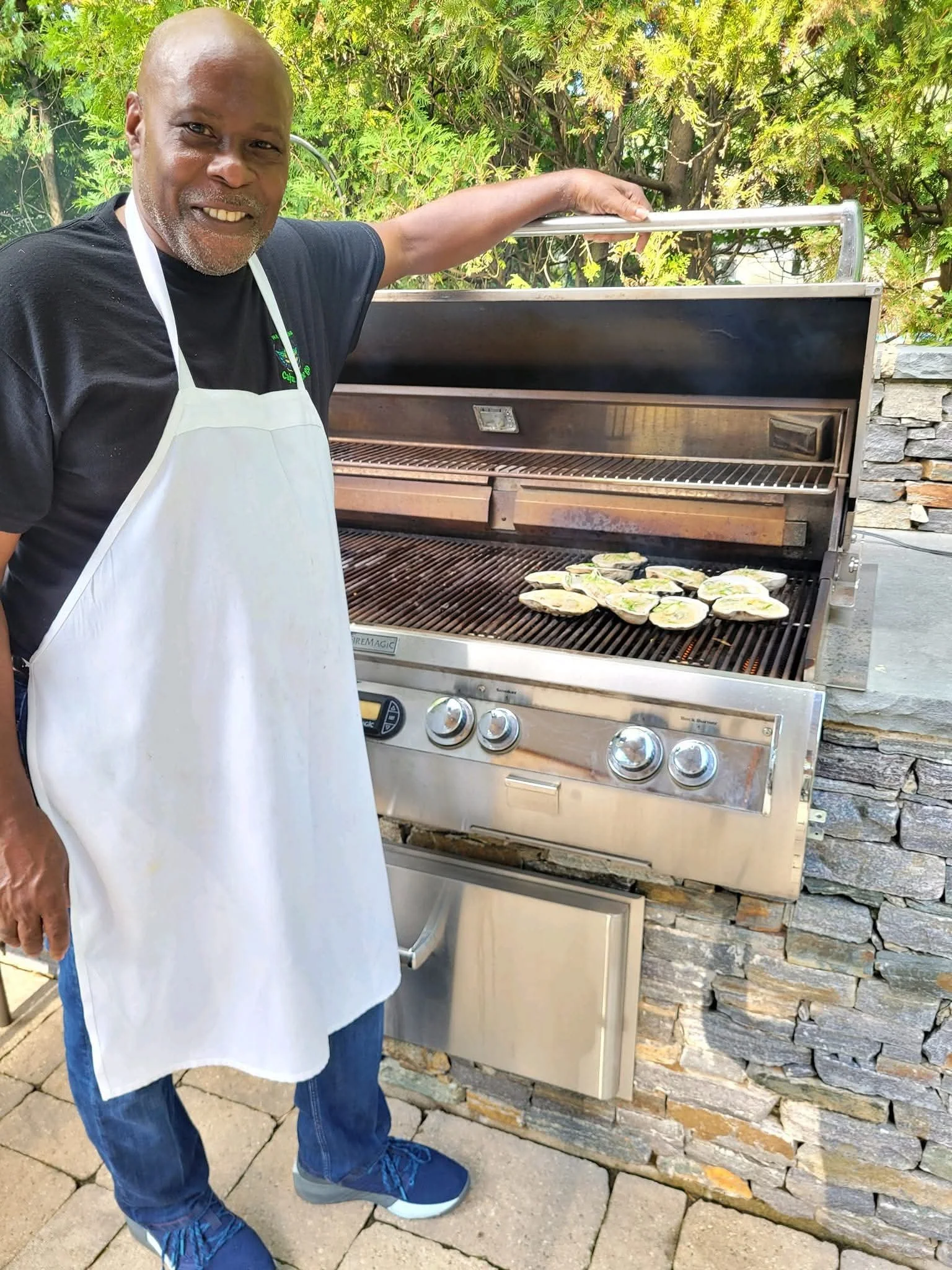 Chef Wyane Hooker wearing a white apron and black t-shirt is grilling small sandwiches on a barbecue grill outdoors, with greenery and stacked stones in the background.